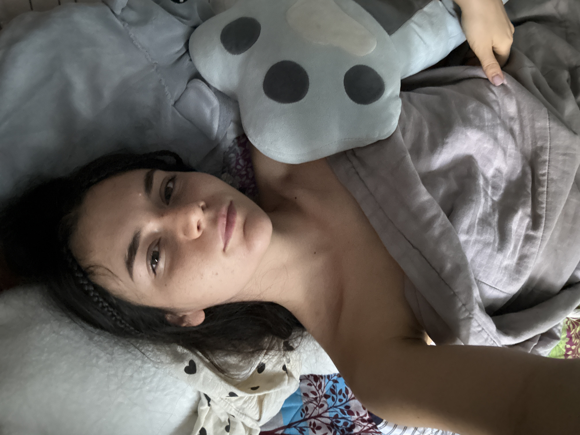 A woman lies on her side on a bed. She has dark hair, light skin, and is covered by a grey blanket. A stuffed animal rests on her chest. The bedding beneath the blanket is patterned. Her head is on a white pillow with black hearts. She appears to be indoors, likely in a bedroom. Location information is not discernible from the image.