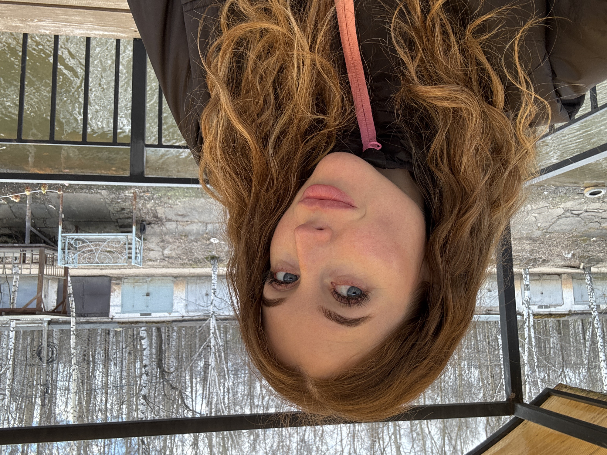 A woman with long, wavy reddish-brown hair is seen from a close-up, upside-down perspective. She has fair skin, blue eyes, and full lips. She is wearing a dark jacket with a pink zipper pull. Behind her, the background appears to be a cityscape, possibly including trees and buildings, reflected in a body of water. The scene is viewed through a metal railing. Based on the available information the image was taken in Chaikovsky, Russia.