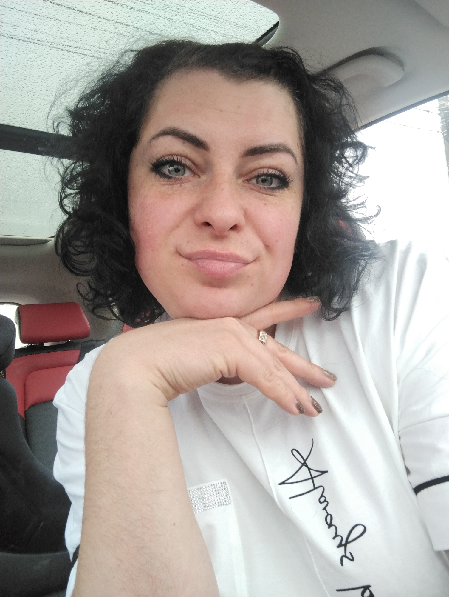 A woman poses for a medium-shot selfie from inside a car, likely in Izhevsk, Russia, on an overcast or rainy day. She has dark, curly hair and light-colored eyes, with a slight, confident smile. Her chin rests on her right hand, which displays a ring with multiple small stones on her ring finger and glittery brown nail polish. She wears a white top featuring a rectangular embellishment of small, square rhinestones on the chest area and stylized black cursive text, possibly "Aramaz," printed on the right sleeve.
The background reveals the interior of the car, including a red and black seat and headrest to her left. Above her, a car window or sunroof shows an exterior view of a dull, grey sky with visible raindrops on the glass, suggesting wet weather. The overall mood conveyed is relaxed and casual.
