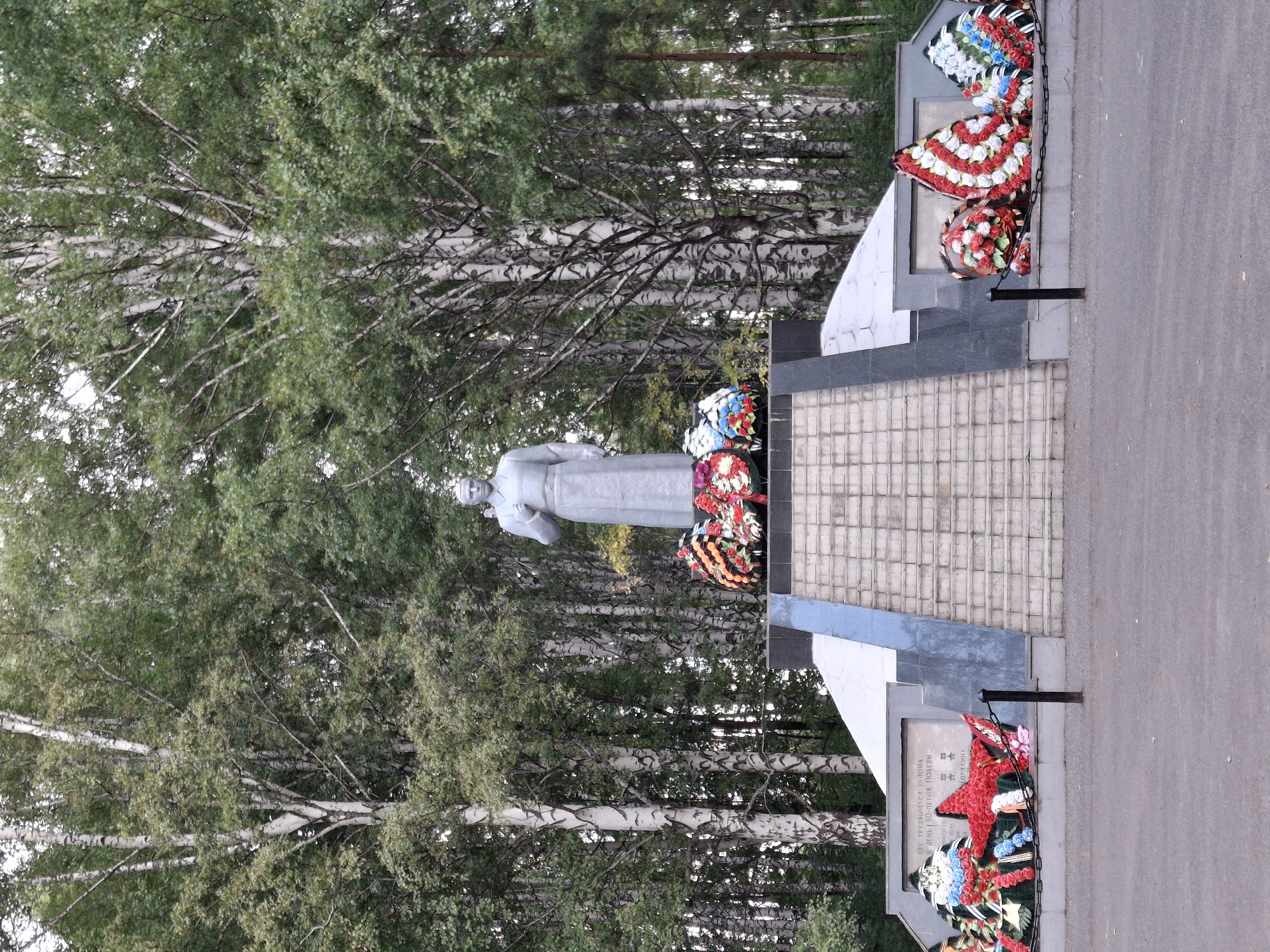 The image shows a monument located in the city of Elovo, Russia. The monument features a central statue of a man in a light-colored robe, standing on a tiered base. The base is adorned with several large, circular wreaths made of red, white, and blue flowers. Steps lead up to the monument. Behind the monument, a dense forest of birch trees with white trunks and green foliage forms the backdrop. The ground in front of the monument is paved. Several black metal posts and chains are placed in front of the monument, marking a boundary. One of the wreaths placed at the base of the monument is shaped like a red star. A plaque is visible on the left side of the monument base, with text in Russian. The sky is overcast.