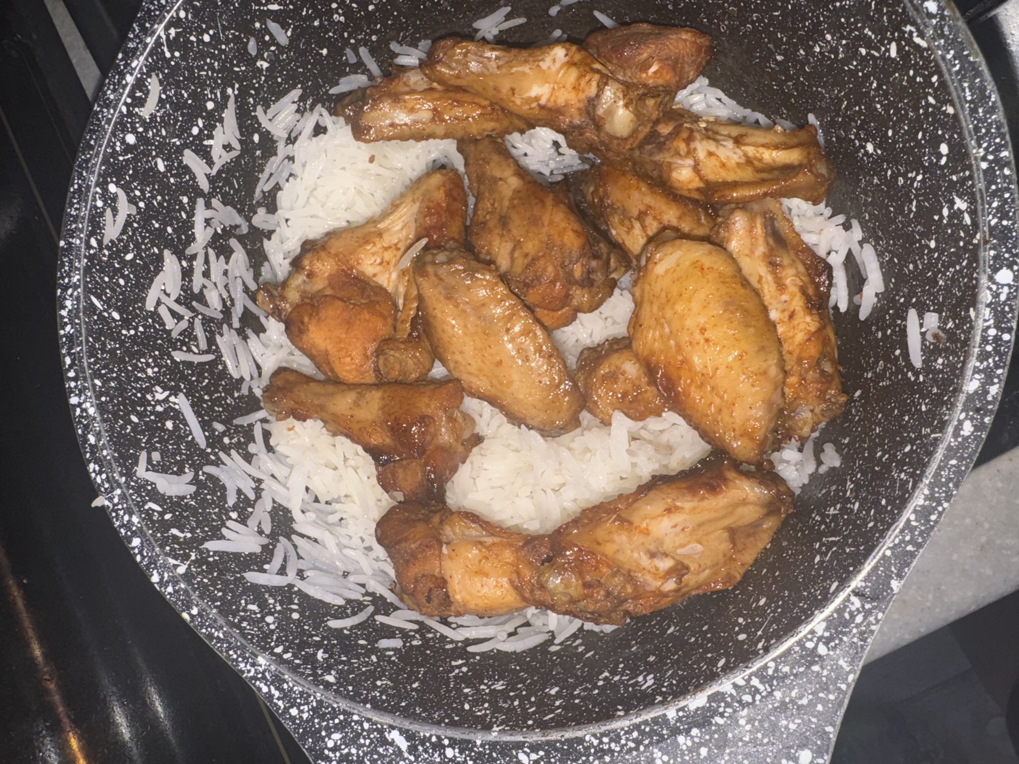 In a black speckled pot, a pile of white rice is arranged in the center, with several glazed chicken wings placed on top. The chicken wings are a deep golden brown and appear to be coated in a glossy sauce. The pot is filled with rice to the brim, and some grains of rice are scattered around the edges of the pot. The background is dark, with a hint of a stovetop visible at the top of the image.
