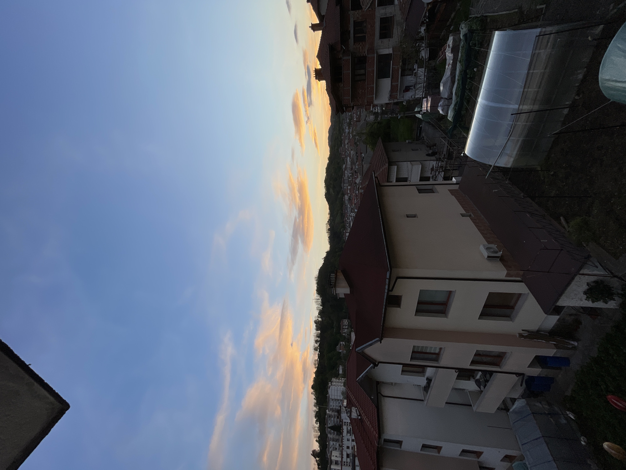 The image depicts a view from an elevated position, possibly a balcony, overlooking a residential area in Zlatograd, Bulgaria. The sky is partially visible, displaying a gradient of blue to orange hues, suggesting either sunset or sunrise. Scattered clouds are present, illuminated by the warm light. The foreground contains rooftops of multi-story buildings with red-tiled roofs and light-colored facades. Several buildings have balconies and windows visible. Further into the scene, a dense area of trees partially obscures a cluster of smaller buildings, likely the town's lower-lying areas. In the background, a partially constructed brick building is visible. A greenhouse is seen in the upper right corner of the image. No people are immediately discernible. The overall scene presents a typical residential setting within the Bulgarian town of Zlatograd, captured during a time of day with soft, warm light.