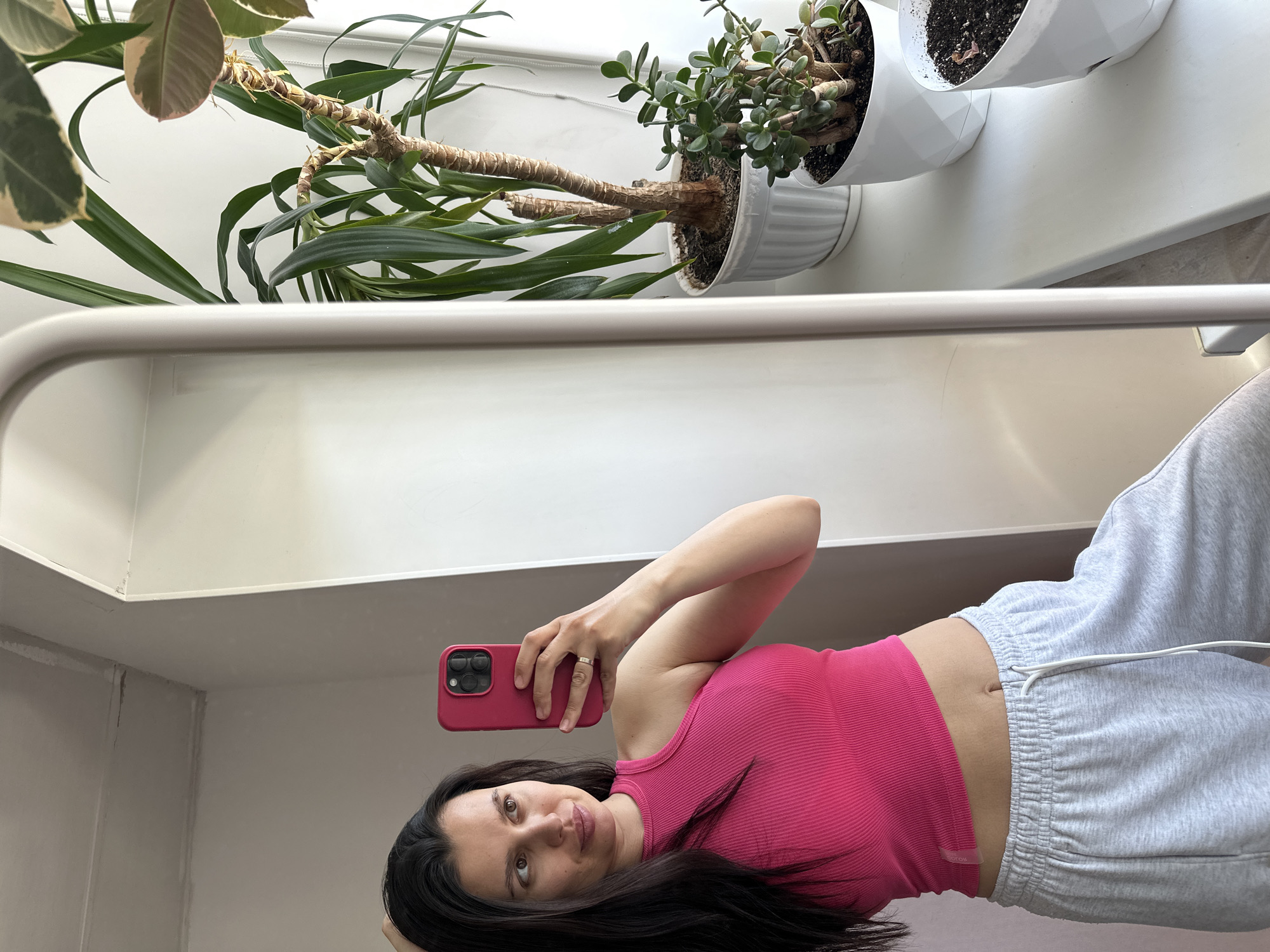A selfie depicts a woman in a pink tank top and grey sweatpants holding a pink smartphone. She is positioned in front of a window ledge displaying three potted plants. The location appears to be indoors. The city is Chaikovsky, Russia.