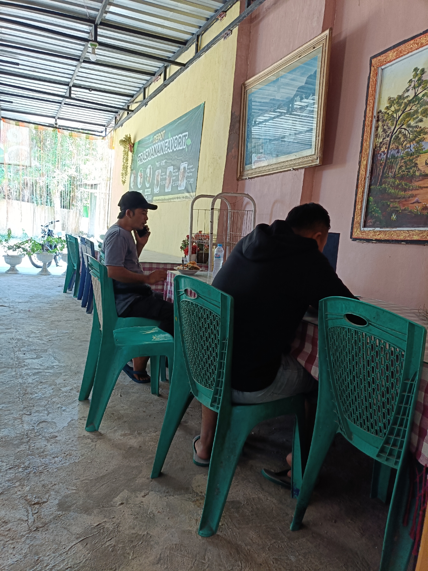 This image depicts an outdoor dining area at Depot Prasmanan Favorit in Pordapor, Indonesia. Two men are seated at tables covered with red and white checkered tablecloths. The man on the left, wearing a grey t-shirt and black shorts, is sitting in a green plastic chair and speaking on his mobile phone. He is facing a table with a plate of food and a bottle of water. The man on the right, wearing a black hoodie and grey shorts, is seated in a green plastic chair and appears to be looking at a laptop. In the background, a banner with the text "DEPOT PRASMANAN FAVORIT" is visible on a light yellow wall. Two framed paintings hang on the pink-toned wall to the right. The floor is concrete, and a line of green plastic chairs extends into the background. Plants and a motorcycle are visible outside the covered area.