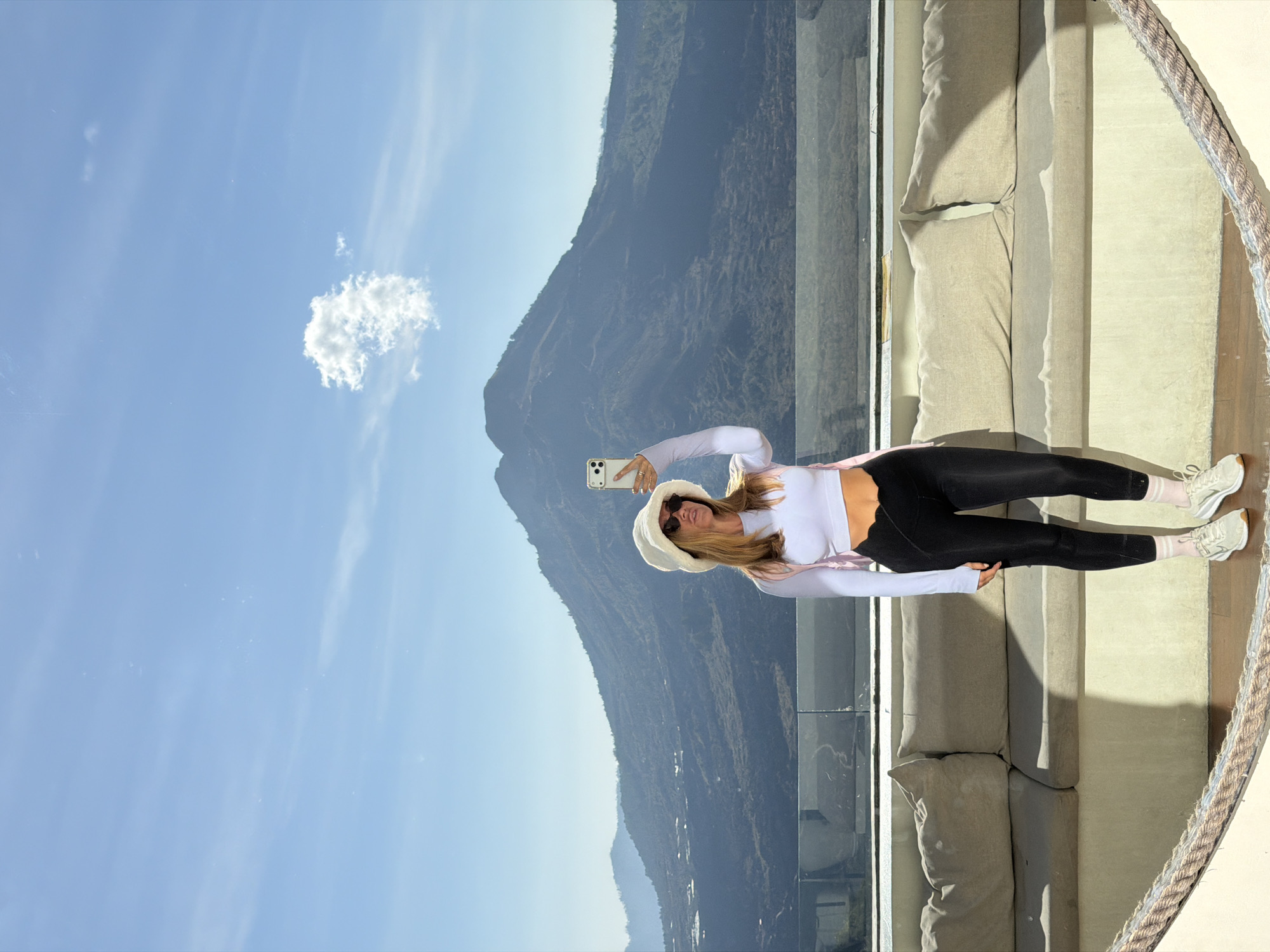 In Kintamani, Indonesia, a woman with blonde hair stands on a balcony, taking a selfie with her phone. She is wearing a white long-sleeved crop top, black leggings, a white bucket hat, sunglasses, and white sneakers with pink socks. A pink strap is visible over her shoulder. She is positioned in front of a large, dark mountain that dominates the background, with a clear blue sky above featuring a small, fluffy cloud. Below the mountain, the landscape appears rugged and undeveloped. In the foreground, a beige sectional couch with large cushions is visible, and a thick rope railing separates the balcony from the viewer. The lighting suggests it is daytime.