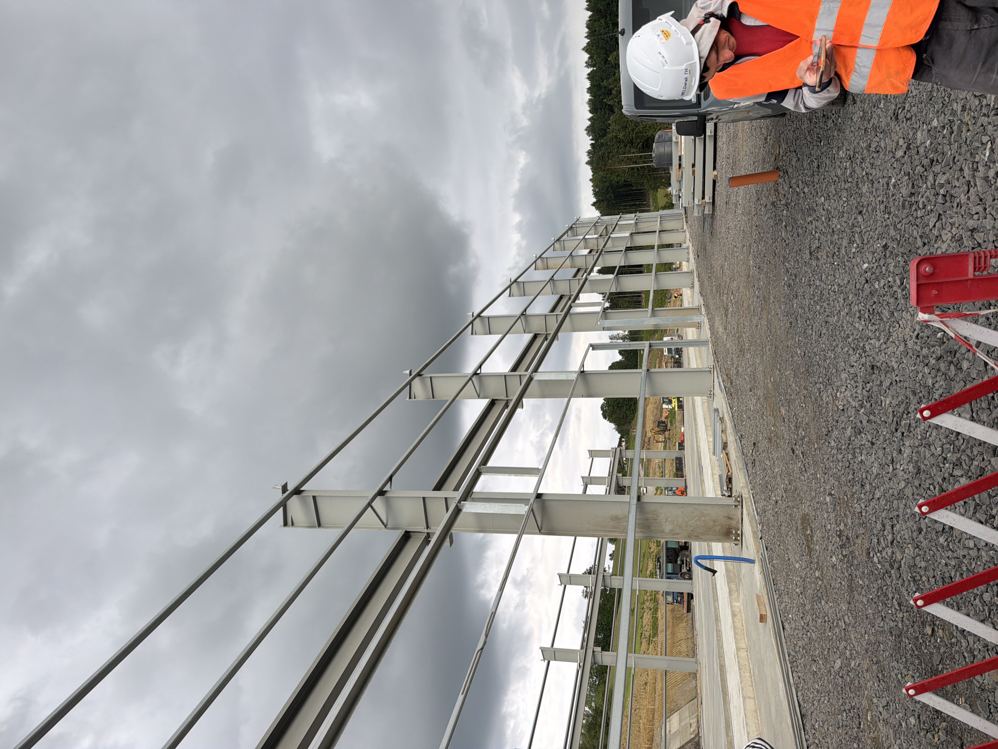 A man wearing a white hard hat and an orange safety vest stands on a gravel surface, looking at his phone. He is partially obscured by a dark gray vehicle to his right. Behind him, a large, unfinished steel structure with visible beams and columns dominates the frame. The sky is overcast with dark clouds. In the background, there is a wooded area with a line of trees and a construction site with heavy machinery, including trucks and excavators. The image appears to have been taken outdoors in Stróża, Poland, as indicated by the context.