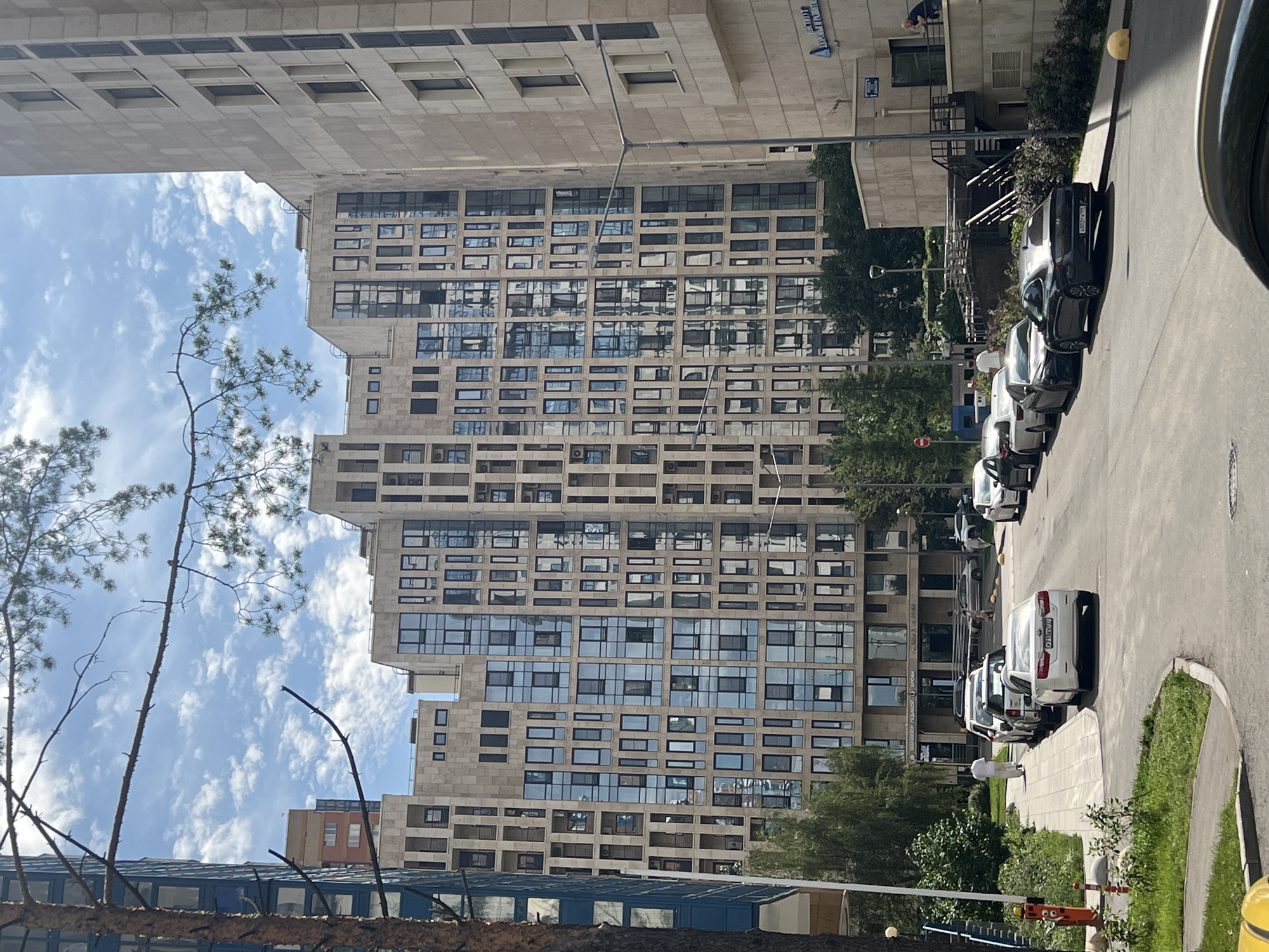 A view of a street in Moscow, Russia, shows a modern residential building with a beige facade and numerous glass windows. A narrow branch from a pine tree hangs in the foreground, partially obscuring the sky. Several cars are parked along the side of the street. A person in a white shirt and pants walks on a paved sidewalk. In the background, a tall building with a blue facade can be seen. The sky is blue with scattered white clouds.