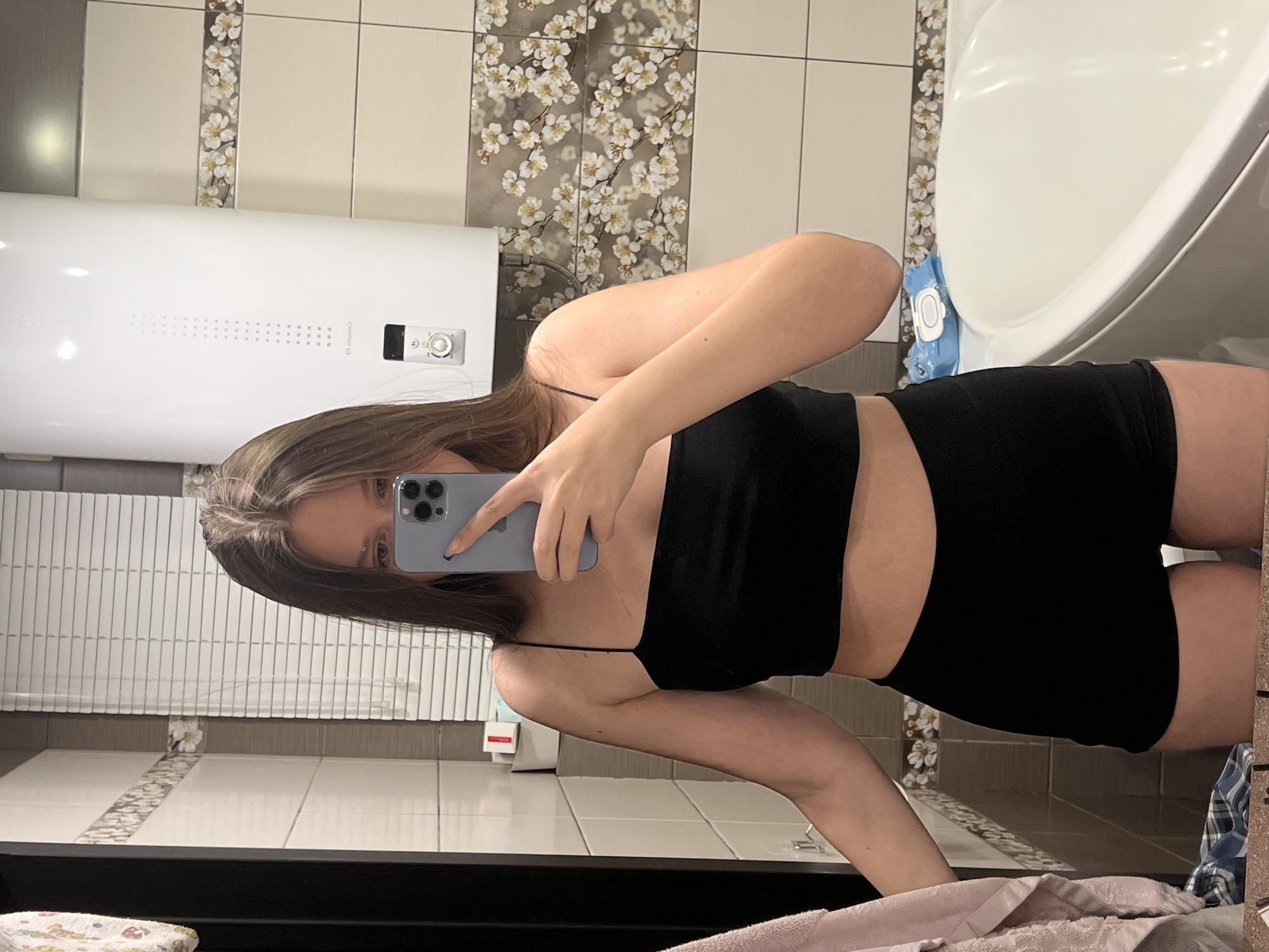 A woman with long brown hair is taking a selfie in a bathroom. She is wearing a black crop top and black shorts. She is holding a grey iPhone in front of her face. The bathroom has white and grey tiles, a white bathtub, a white water heater, and a floral decorative border on the tiles. The location is potentially Chaikovsky, Russia.