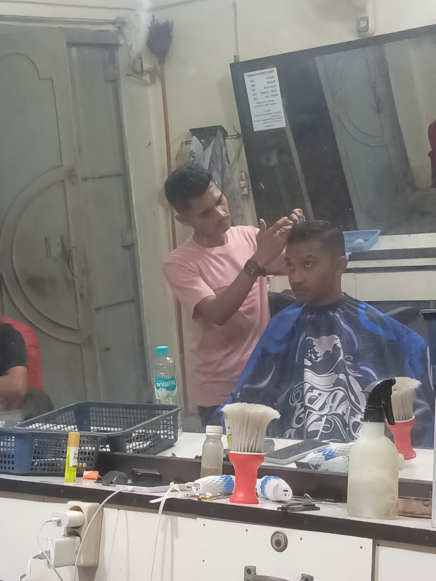 The image depicts a barber shop, likely located in Lhokseumawe, Indonesia, with two men present. One man, wearing a pink t-shirt, is actively cutting the hair of the other man, who is seated and wearing a blue and black barber cape. The barber is using his hands to style the seated man's hair.
The environment appears to be a well-lit indoor space, possibly with a mirror reflecting some of the scene. On a counter or shelf in the foreground, various barber tools and supplies are visible. These include a black basket filled with items, a bottle of water with a blue cap, a spray bottle, two brushes with red bases, and several other unidentifiable objects. Power outlets with charging adapters are also present on the wall in the foreground. The overall impression is of an active and functional barber shop.