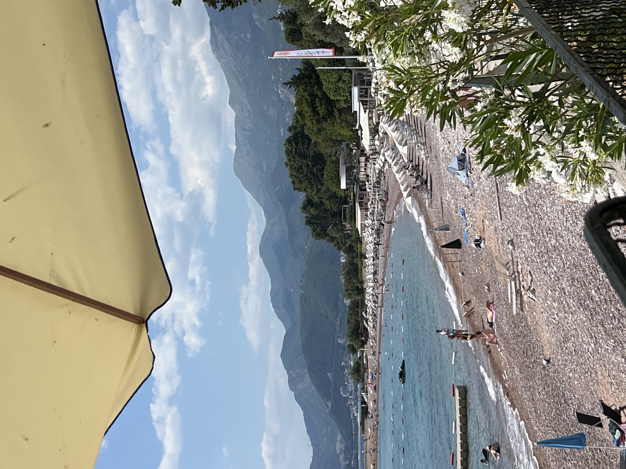 This is a vertically oriented, wide-angle photograph of a crowded beach scene under a clear blue sky with scattered white clouds. The sun is bright, suggesting it is daytime. The beach is covered in pebbles, and rows of lounge chairs with white umbrellas are set up along the shore. The turquoise water of the sea gently laps at the beach, with some people swimming and wading.
In the foreground, a large portion of a yellow beach umbrella is visible on the left side of the frame, providing shade. Beyond the chairs, a line of trees separates the beach from a backdrop of imposing, green-covered mountains that rise steeply into the sky. A small flag with red and white markings is planted on a pole near the treeline, and some buildings are visible nestled between the trees and the mountains.
Further down the beach, small groups of people are seen relaxing on lounge chairs, walking along the shore, and swimming. A few boats or inflatable watercraft are present in the water. The overall atmosphere is one of leisure and recreation on a sunny day at a popular seaside resort.
There is some text visible on a small, rectangular flag on a pole. It appears to be stylized lettering in red and white, but it is too small and distant to be legible. The scene evokes a feeling of a summer vacation.
