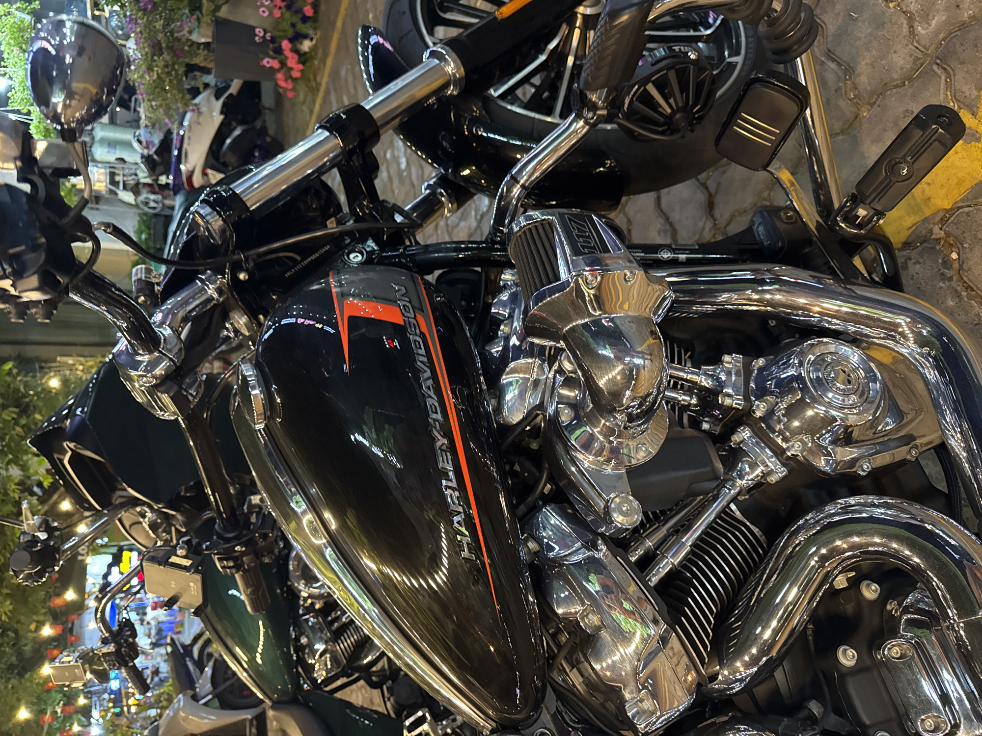 The image features a close-up of a Harley-Davidson motorcycle, focusing on the engine and fuel tank. The tank is black with an orange stripe and the Harley-Davidson logo. The engine is highly polished chrome. The background is blurred but appears to show a city street scene with flowers and other motorcycles. The location cannot be determined to be Đà Nẵng, Vietnam based on the provided information.
