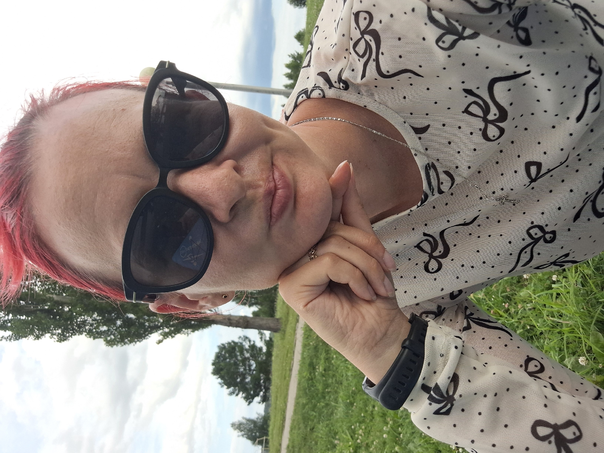 A close-up, high-angle selfie is taken by a woman with bright pink hair. She is outdoors, wearing black, oversized sunglasses and a white, sheer long-sleeved shirt with a pattern of black dots and bows. Her right hand is resting on her chin, adorned with a ring on her index finger. She is wearing a black smartwatch on her left wrist. The background shows a grassy area with a path, trees, and a cloudy sky. There is no indication of the city of Atamanovka, Russia.