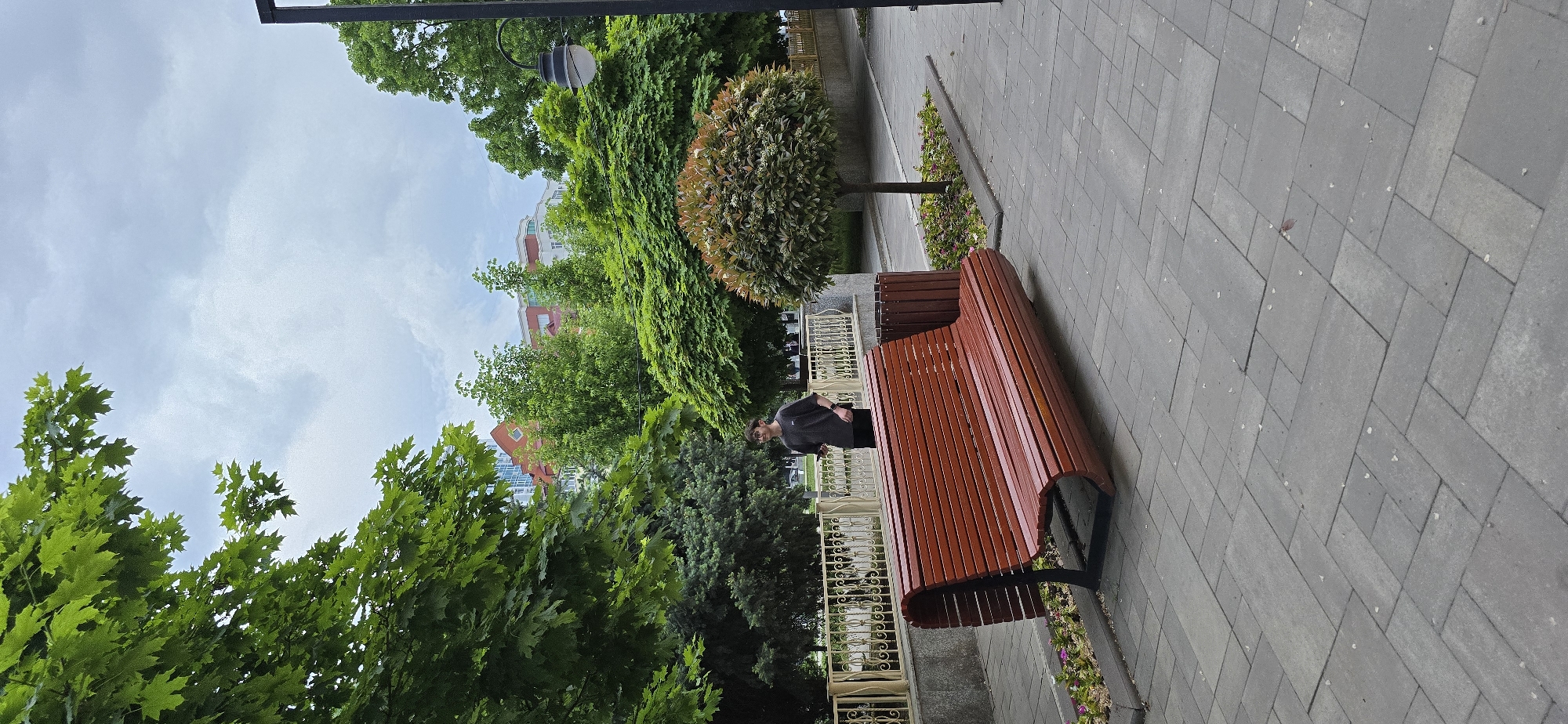 A horizontal shot of a scene in Grozny, Russia. A fair-skinned young man with glasses stands near a modern, curved, reddish-brown wooden bench on a gray-paved sidewalk with light trees, bushes, and buildings behind him. The man is wearing a black t-shirt and black pants. The background features a wrought iron fence, green trees, a circular bush, and buildings with light blue skies. The scene is taken during the day, with overcast lighting. A black pole and light fixture are in the top left corner.