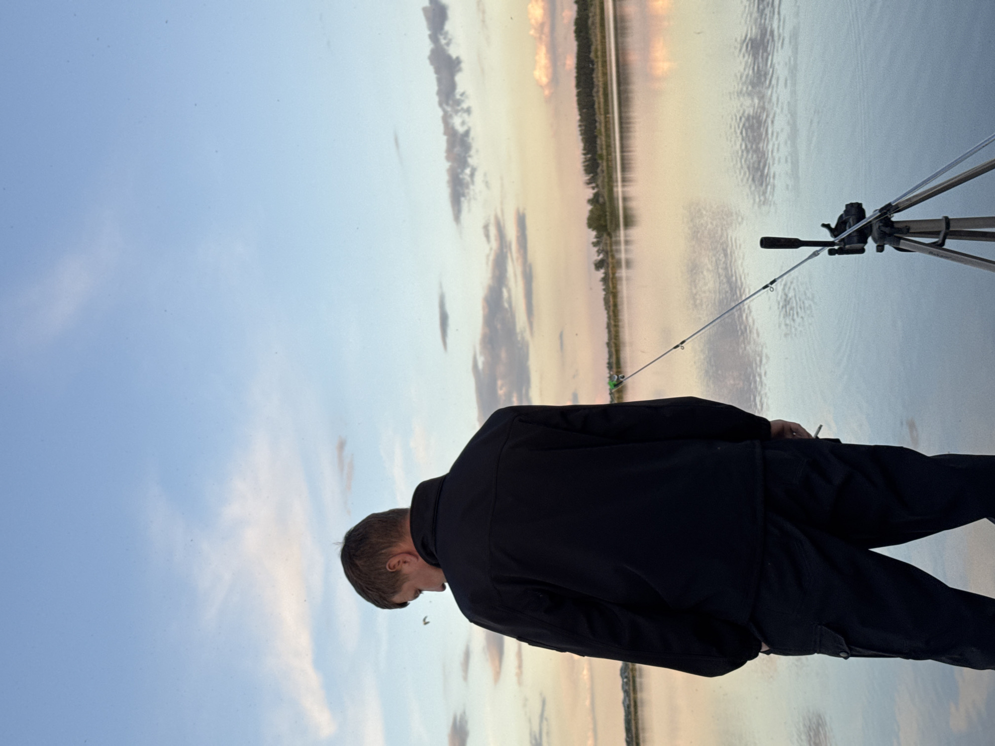 A man in a black jacket and pants is standing by a lake in Unknown, Unknown. He is holding a fishing rod, which is attached to a tripod. The sky is blue with some clouds, and the sun is setting over the lake. The reflection of the sky is visible on the surface of the water. There are trees in the background.