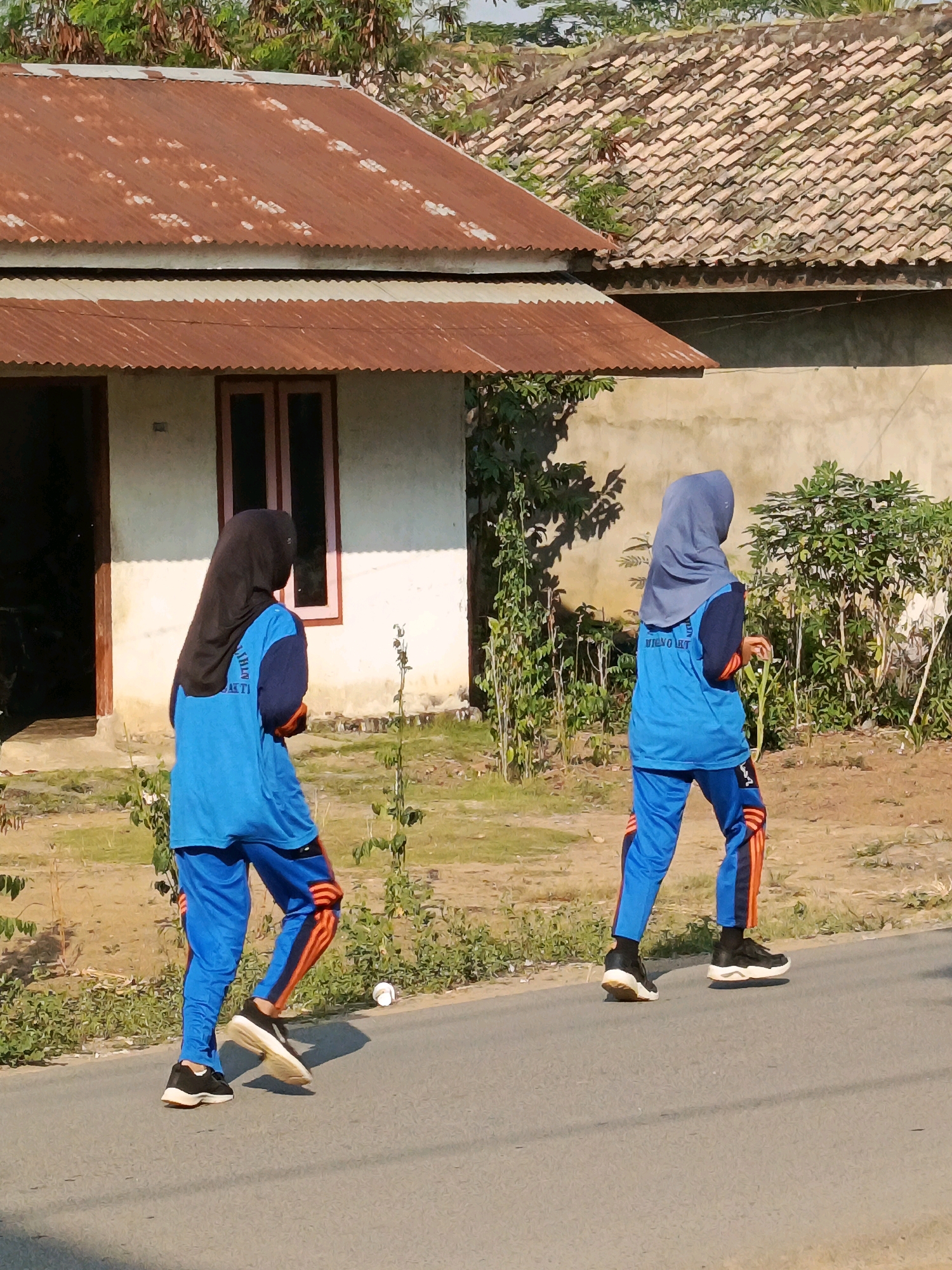 In Megang Sakti I, Indonesia, two individuals wearing athletic attire are captured walking on an asphalt road. The person on the left wears a black hijab and a blue long-sleeved shirt with blue pants featuring orange stripes. The person on the right wears a grey hijab, a blue long-sleeved shirt, and blue pants with orange stripes. Their outfits suggest they are participating in some form of physical activity.
Behind them, a building with a rusty, corrugated metal roof and a tiled roof can be seen. The building has a white exterior wall with a pink-framed window. Vegetation, including bushes and trees, surrounds the building and the roadside, indicating a semi-rural or residential area. The lighting suggests it is daytime, with shadows cast on the road and ground. The overall scene depicts a casual outdoor activity in a residential environment.