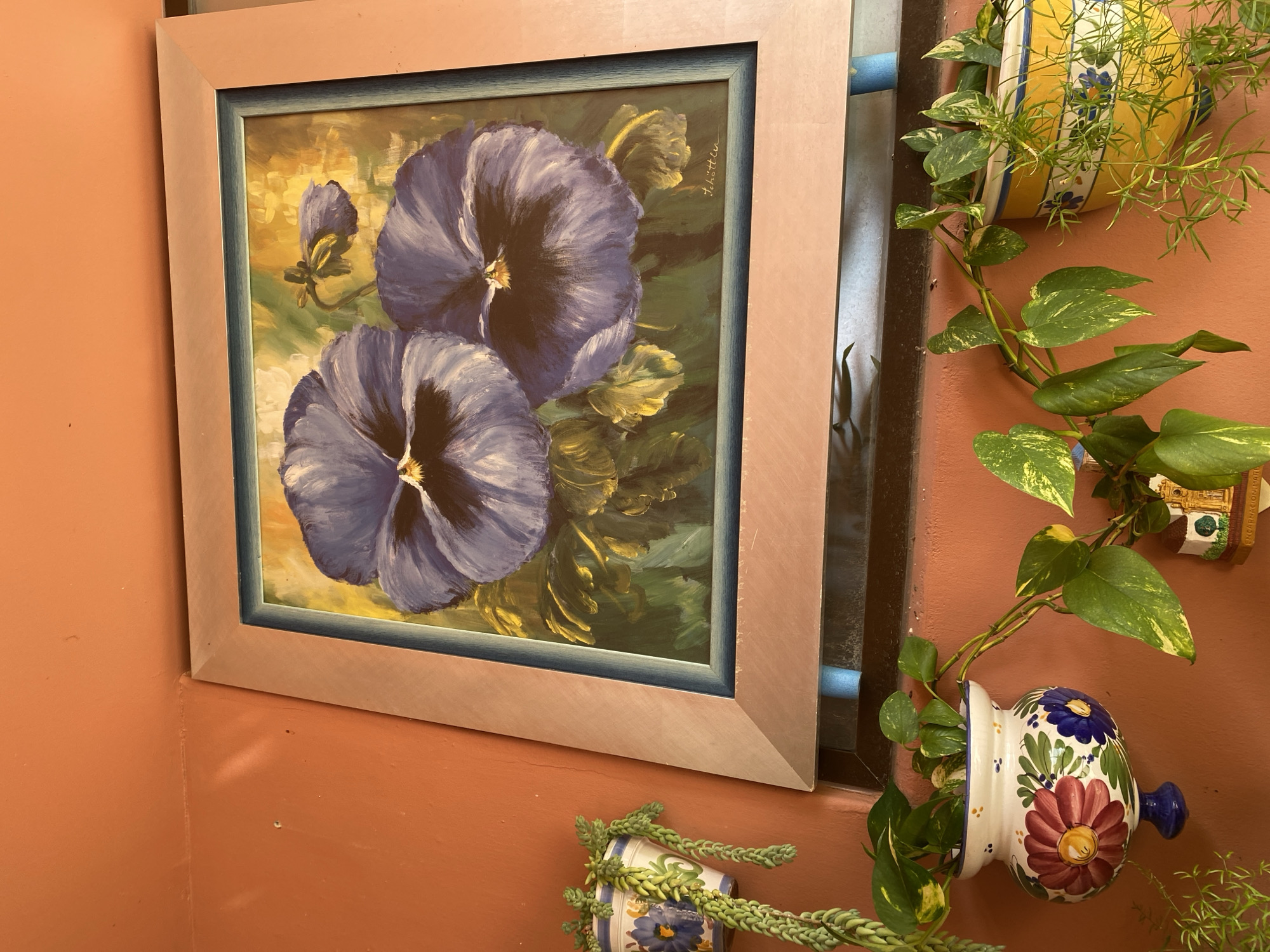A framed painting of two purple pansies is displayed on an orange wall. The painting, which appears to be oil on canvas, has a light pink frame and a blue inner matting. A signature, "Schöttler," is visible in the upper right corner of the painting. Hanging plants are present on either side of the painting. To the right, a vibrant yellow ceramic pot, decorated with blue flowers, holds a trailing plant. Below this, another ceramic planter, adorned with painted flowers in red, blue, and yellow, also supports a trailing plant. To the left of the painting, a thin, light-colored vertical element is visible. The overall impression is of an outdoor or semi-outdoor space, possibly a patio or balcony, decorated with art and plants. The lighting suggests daytime.