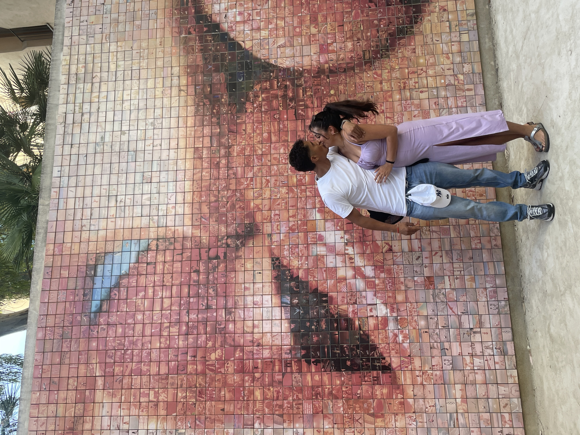 This image captures a young adult male and female embracing and kissing in front of a large mosaic mural in Plaça d'Isidre Nonell, Barcelona, Spain. The mural, known as "The Kiss of Freedom" (El Petó), is constructed from numerous individual photographic tiles, which coalesce to form a detailed close-up of a human eye with a blue iris and a portion of a face, predominantly in shades of pink and red.
The male, positioned on the left, has medium-brown skin and short, dark, curly hair. He wears a white short-sleeved t-shirt, light blue denim jeans, and dark athletic shoes with white accents. A white baseball cap with a dark "NY" logo is clipped to his right hip pocket, and a black backpack strap is visible over his left shoulder. His right arm is around the female's waist as he leans in. The female, positioned on the right, also has medium-brown skin and dark hair styled in a ponytail. She wears a light purple, strappy midi-dress with a high slit on the left leg, silver flat sandals, and small earrings. Her left arm is around the male's neck, and her right hand rests on his abdomen. Both individuals are engaged in a kiss. The ground is light grey concrete pavement, and above the mural, green palm fronds and a section of a light-colored building are visible against a bright sky.