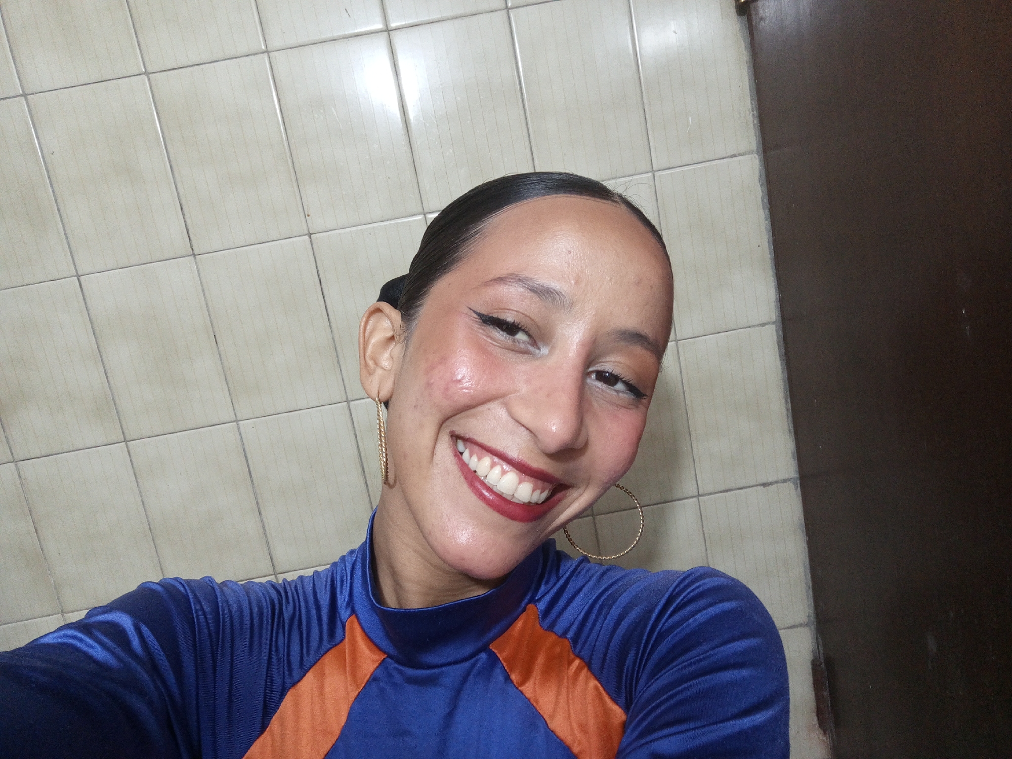 A person with dark hair pulled back in a bun is smiling directly at the camera. She is wearing large gold hoop earrings and a blue and orange long-sleeved shirt. The background is a wall tiled in light beige squares and a dark brown door. The location is likely a bathroom within a building, but there is no definitive evidence to connect it directly to Porlamar, Venezuela.