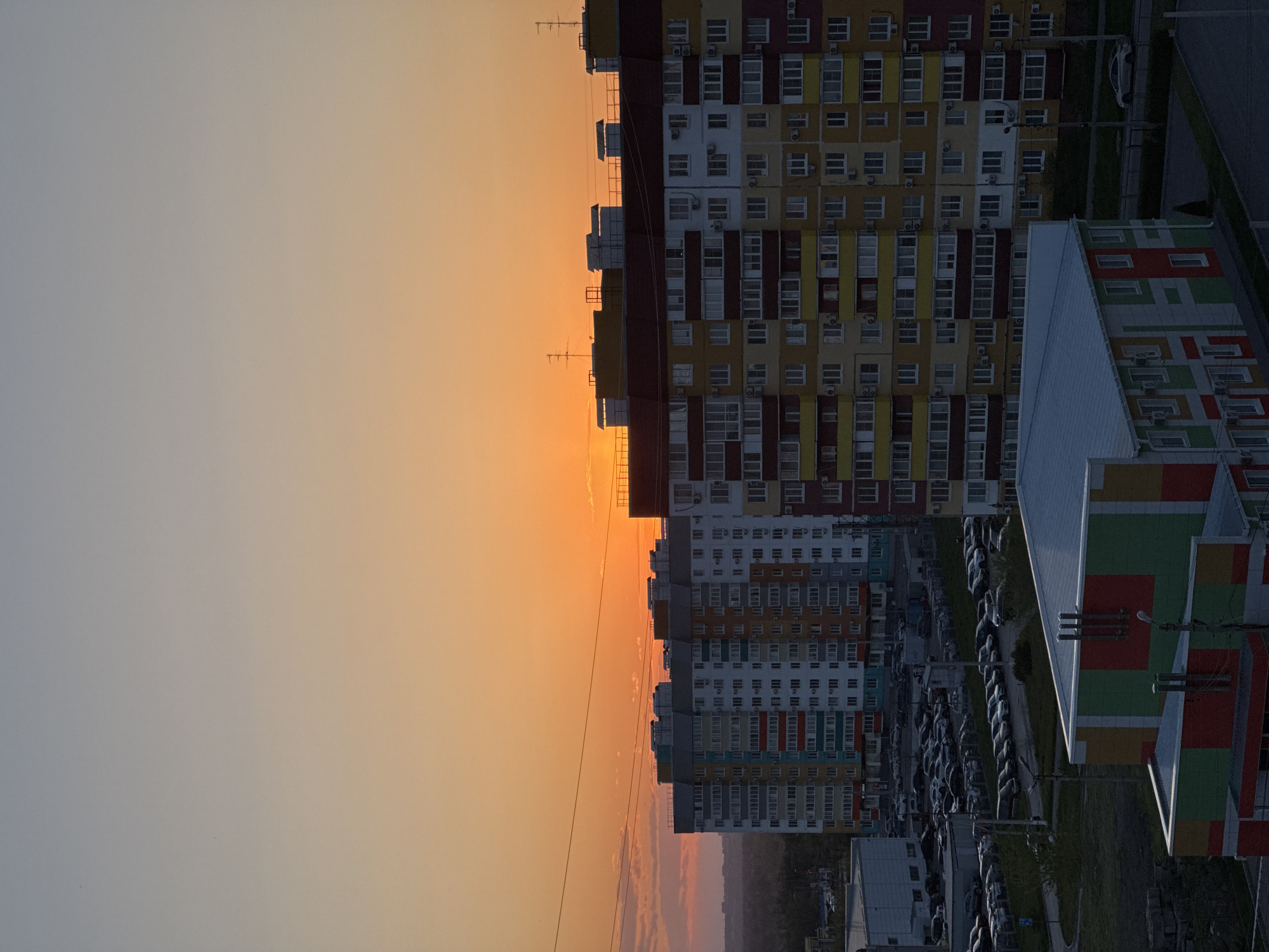 The image presents an urban landscape captured during sunset. The sky, which dominates the upper two-thirds of the frame, exhibits a striking gradient, transitioning from a warm, intense orange-yellow glow on the left horizon, indicating the position of the setting sun, to a soft, hazy grey-blue towards the upper right.
Stretching across the mid-ground and right side of the image is a dense collection of multi-story residential buildings. These modern apartment blocks are characterized by their colorful facades, featuring a vibrant mix of yellow, red, white, blue, and green panels in a patchwork style. Numerous windows and balconies are visible on their exteriors, alongside external air conditioning units. Some rooftops are adorned with antennas, slightly silhouetted against the bright sky.
In the foreground, on the lower right, a distinct building with a light, sloped roof stands out. Its visible side features a bold, geometric pattern composed of green, red, yellow, and white sections. Below the residential structures, extensive parking lots are visible, densely packed with numerous parked cars. Paved roads, pathways, and some patches of green grass or low vegetation complete the ground level. Utility poles and streetlights are scattered throughout the scene.
No individuals are visible, suggesting a focus on the architectural and environmental elements of the city at dusk. The vibrant building colors contrast with the natural hues of the sunset sky, depicting a bustling, modern residential district in Volzhsky, Russia. There is no discernible text in the image.