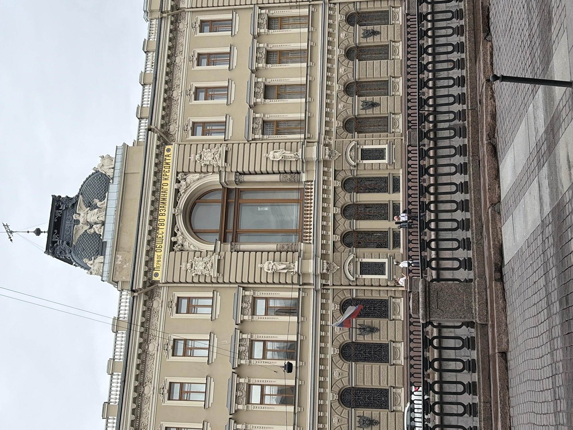 The image shows a section of the facade of a historic building in Saint Petersburg, Russia. The building features classical architectural elements such as ornate cornices, pilasters, decorative sculptures, and arched windows with intricate metalwork. Above a prominent central arched window, there is a golden inscription that reads "ПЕРВОЕ ОБЩЕСТВО ВЗАИМНАГО КРЕДИТА," translating to "First Mutual Credit Society." The building's facade is a light beige color, with contrasting darker tones on the roof and window frames.
In the foreground, there is a black metal railing running along a canal or embankment. Beyond the railing, the building's lower level is visible, with more arched windows and decorative elements. A Russian flag is prominently displayed on the building's facade, slightly to the left of center. Several people are visible walking along the embankment, and a white car is parked on the left side of the image. The ground in the immediate foreground is paved with cobblestones. The sky is overcast, suggesting a cloudy day. The overall impression is of a grand, well-preserved historical building in an urban setting.