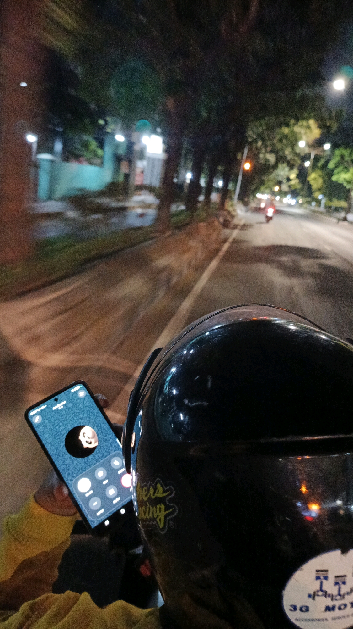 The image depicts a first-person perspective of someone riding a motorcycle at night in Surakarta, Indonesia. The rider is holding a smartphone, which displays a video call interface with a person's face visible in a circular frame. The background shows a street scene with trees lining the road, streetlights, and other vehicles, including a motorcycle in the distance. The overall atmosphere suggests a typical urban night setting.