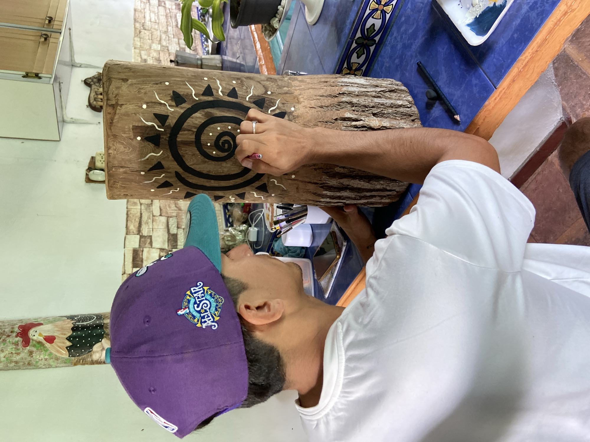 The image captures a person, likely male, from a top-down and profile perspective, engrossed in painting a wooden log. He wears a white t-shirt and a purple and teal baseball cap, which prominently displays "ALL-STAR" within a decorative emblem featuring a globe and stars, along with a small "NBA" logo. His right hand, with a silver ring on the ring finger, meticulously applies small white dots using a pink-tipped brush or tool onto a black spiral design on the log.
The natural wooden log, with its textured bark, is adorned with a central black spiral, surrounded by black triangular shapes and white wavy lines and dots. The activity appears to be a focused artistic or crafting session.
The setting is an indoor space, possibly a home or workshop. The log rests on a vibrant blue tiled counter, which also features a decorative patterned tile. Nearby, various art supplies including brushes and a paint palette are visible. In the background, white kitchen cabinets and an earthy-toned tiled floor can be seen. A small, colorful ceramic rooster decoration is affixed to a wall. The overall ambiance suggests a creative and tranquil environment. This scene is set in Los Velásquez, Venezuela, likely during the daytime given the clear ambient lighting.