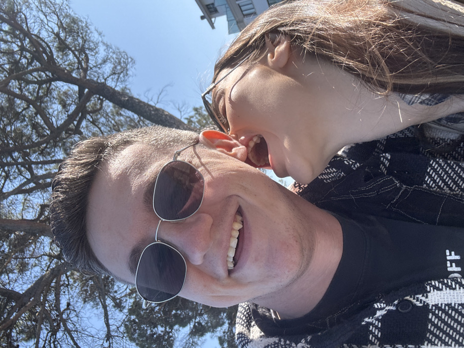 This close-up outdoor photograph depicts two adults engaging in a playful interaction in Peschiera del Garda, Italy. The central figure, an adult male, occupies the left side of the frame, smiling broadly towards the camera, revealing his upper teeth, which include two prominent, elongated canines. He wears silver-rimmed sunglasses with dark lenses and a black and white plaid flannel shirt over a black t-shirt with white lettering on the chest. To his right, an adult female is positioned with her face angled towards the male's right ear, her mouth wide open in an expression suggestive of a laugh or shout. Her long, light brown hair is visible, and eyeglasses are perched atop her head. The background features a large tree with dark, gnarled branches and sparse foliage, set against a clear blue sky. In the distant right background, a light-colored, multi-story building with visible balconies can be discerned, consistent with a lakeside or coastal architectural style. The scene is brightly lit by natural sunlight.
