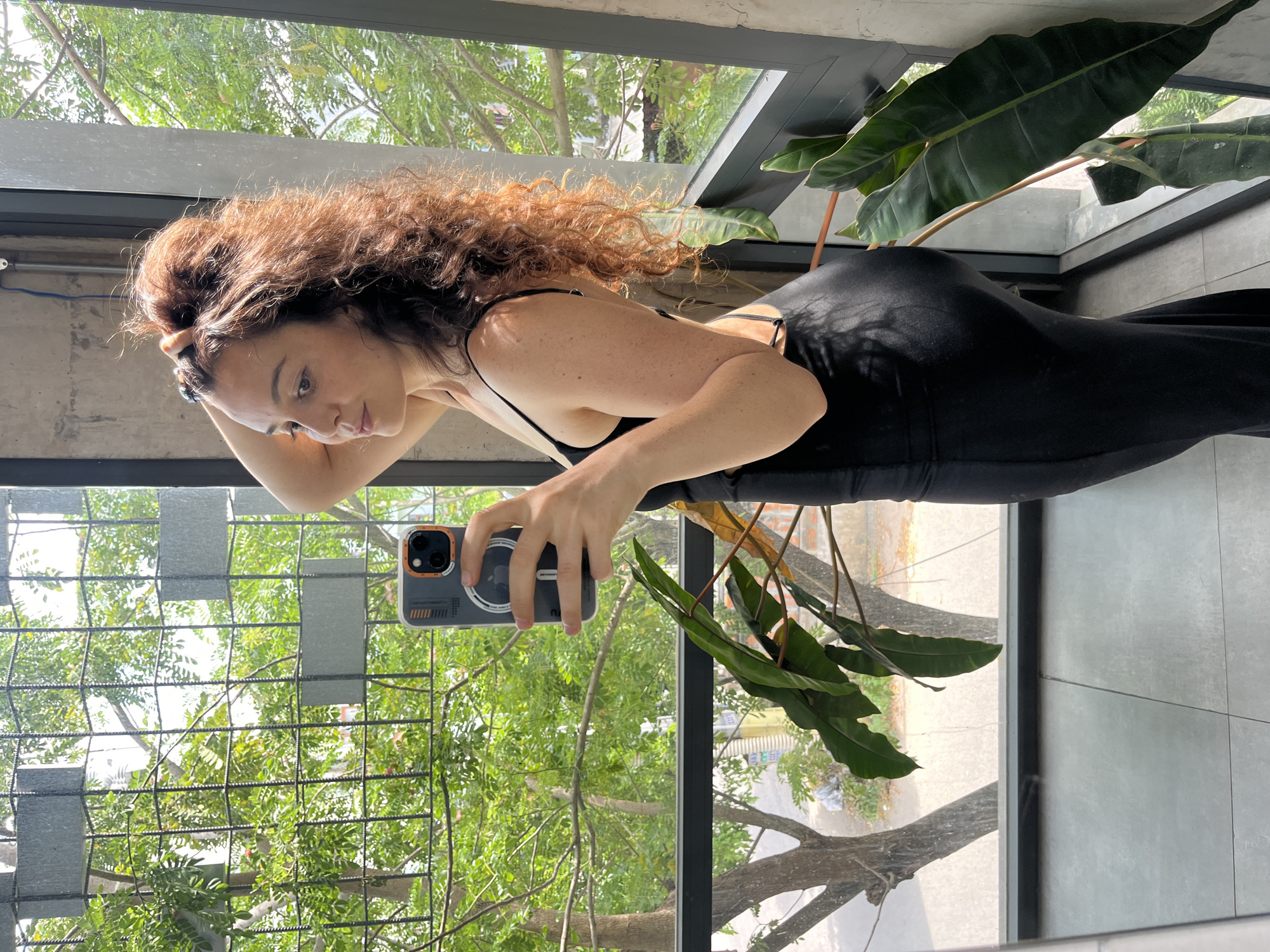 The image shows a young woman with curly brown hair holding a smartphone in her left hand and taking a mirror selfie. She is wearing a black dress with thin straps and an open back. Her right hand is raised, touching her hair. She is standing in front of a large window with a view of trees and foliage. A metal grate is visible on the left side of the window. The flooring is tiled. The background suggests an indoor space with a concrete wall and a dark-framed window. The context of Phu Thuy Ward, Vietnam, is not visually verifiable from the provided image.