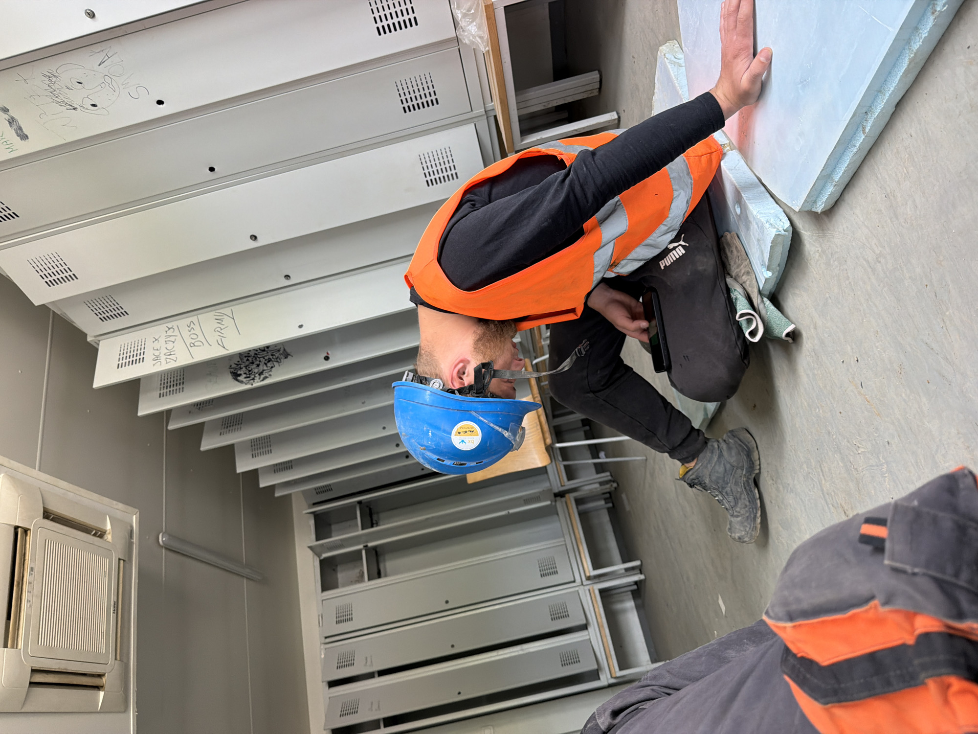 In Męcina, Poland, within a locker room, a man wearing a blue hard hat and an orange high-visibility vest is seen crouching on the floor. He is looking down at a mobile phone in his hand. The environment features rows of grey metal lockers, some open and empty, others closed. A wooden bench runs along the base of the lockers. The floor is concrete. In the foreground, another person's legs, clad in grey work trousers with orange and black stripes, are visible. The overall scene suggests a utilitarian setting, possibly a construction site or industrial facility.