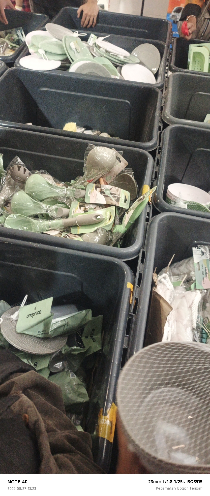 This image shows a close-up, slightly blurry view of several dark gray plastic bins filled with kitchen utensils and tableware. The items are predominantly green and gray, many of them still in their original plastic packaging. Some items appear to be spatulas or serving spoons, while others are plates or bowls. A label that reads "oneprice" is visible on some of the packaging. In the lower portion of the image, a brown fabric or clothing item is partially visible, along with a cylindrical object with a mesh pattern. The bottom of the image contains text indicating "NOTE 40", a date and time "2026.08.27 15:23", and camera settings "23mm f/1.8 1/25s ISO5515" followed by the location "Kecamatan Bogor Tengah". The background is out of focus, suggesting an indoor setting, possibly a retail or storage area.