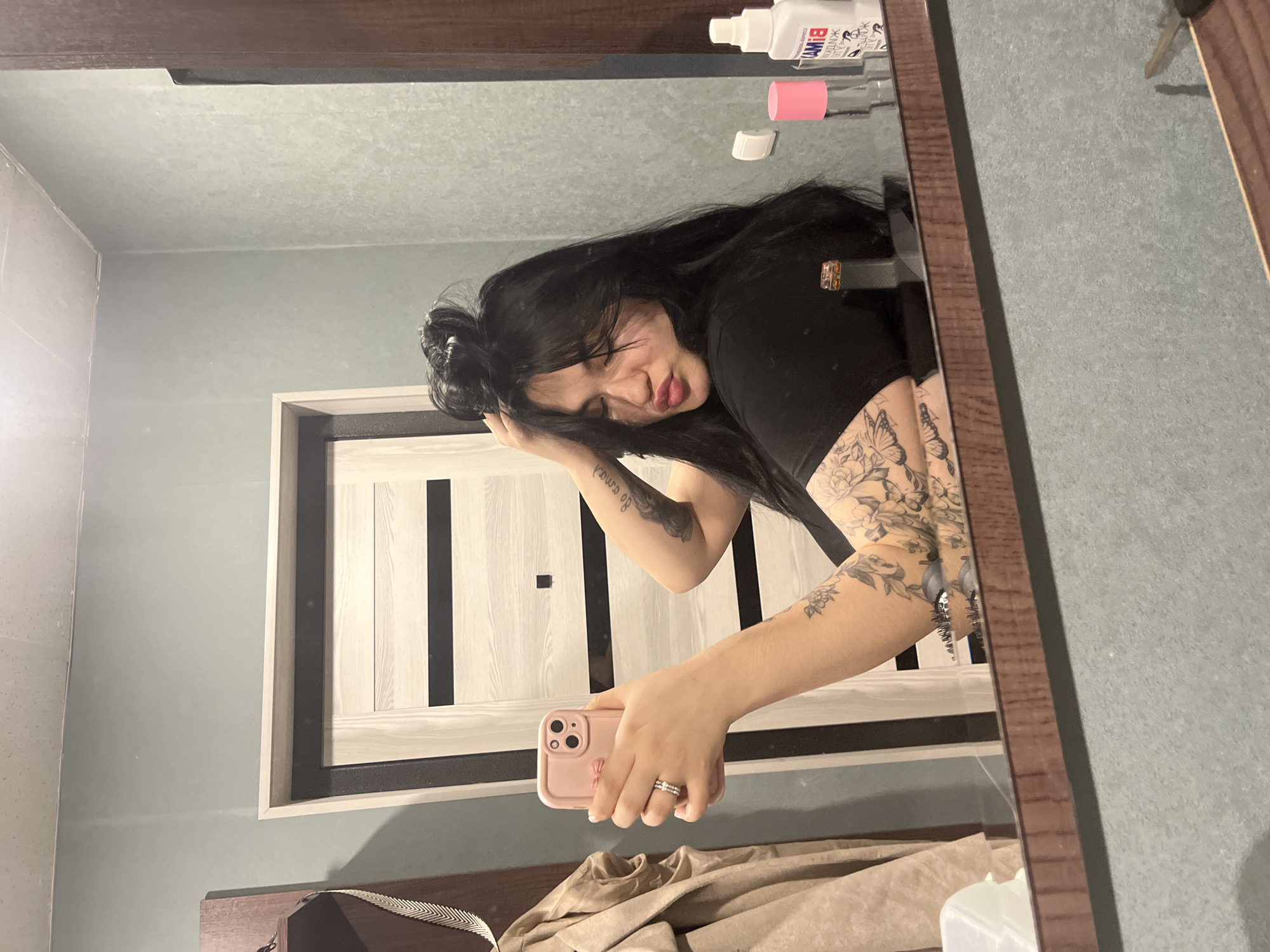 A female subject is captured in a mirror selfie within an indoor residential setting. She is depicted from the chest up, with long, dark hair styled partly in a messy bun, and strands framing her face. Her eyes are partially closed, and her lips are pursed in a "duck face" expression. She wears a dark, short-sleeved top. Tattoos are visible on both arms, featuring floral designs, a butterfly, and script. Her left hand, adorned with a two-band silver ring, holds a pink smartphone with a matching case, pointed towards the mirror. Her right hand is raised, with fingers near her hair. The mirror's reflection reveals light grey-green textured walls and a white ceiling. A light-colored wooden door with two dark horizontal stripes and a black frame is visible in the background. On the surface in front of the mirror, a pink bottle, a white bottle with Russian text, a ring stand holding a gold-colored ring, a white plastic container, and a key are present. To the left, a light-colored jacket or coat hangs on a wooden shelf, from which a silvery decorative strip also dangles. A white light switch or outlet is on the wall. The specific location within Chaikovsky, Russia, is not discernible from the provided visual information.