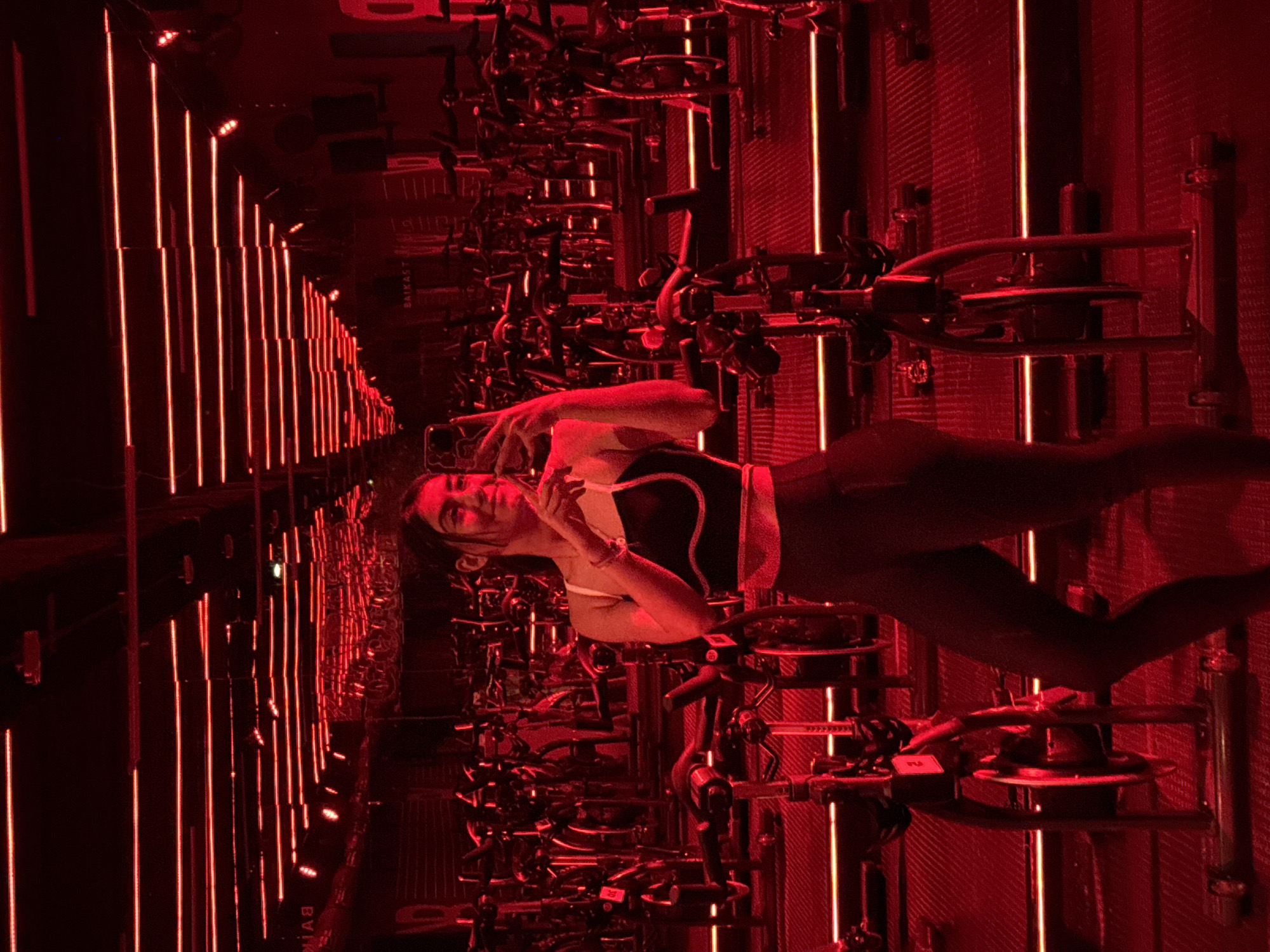 A young woman is seated on an exercise bike in a fitness studio, illuminated by intense red lighting. The studio is filled with rows of exercise bikes. The lighting consists of vertical LED bars and some are installed in the walls with reflective metal panels. The studio is identified as B-CYCLE located in Puebla, Mexico.