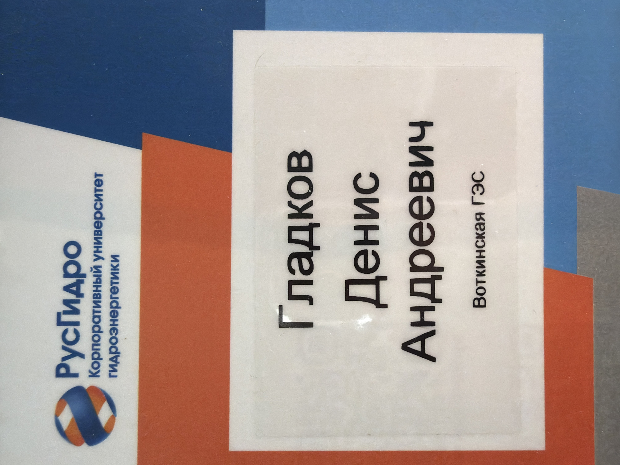The image shows a name tag with the text "Гладков Денис Андреевич" (Gladkov Denis Andreevich) and "Воткинская ГЭС" (Votkinsk HPP). It's likely a name tag identifying a person working at the Votkinsk Hydroelectric Power Plant. The logo of "РусГидро" (RusHydro) and the words "Корпоративный университет гидроэнергетики" (Corporate University of Hydropower Engineering) appear on a card behind the name tag. Votkinsk HPP is located in the city of Chaikovsky, Perm Krai, Russia. The background consists of layered shapes of blue, white, orange, and grey.