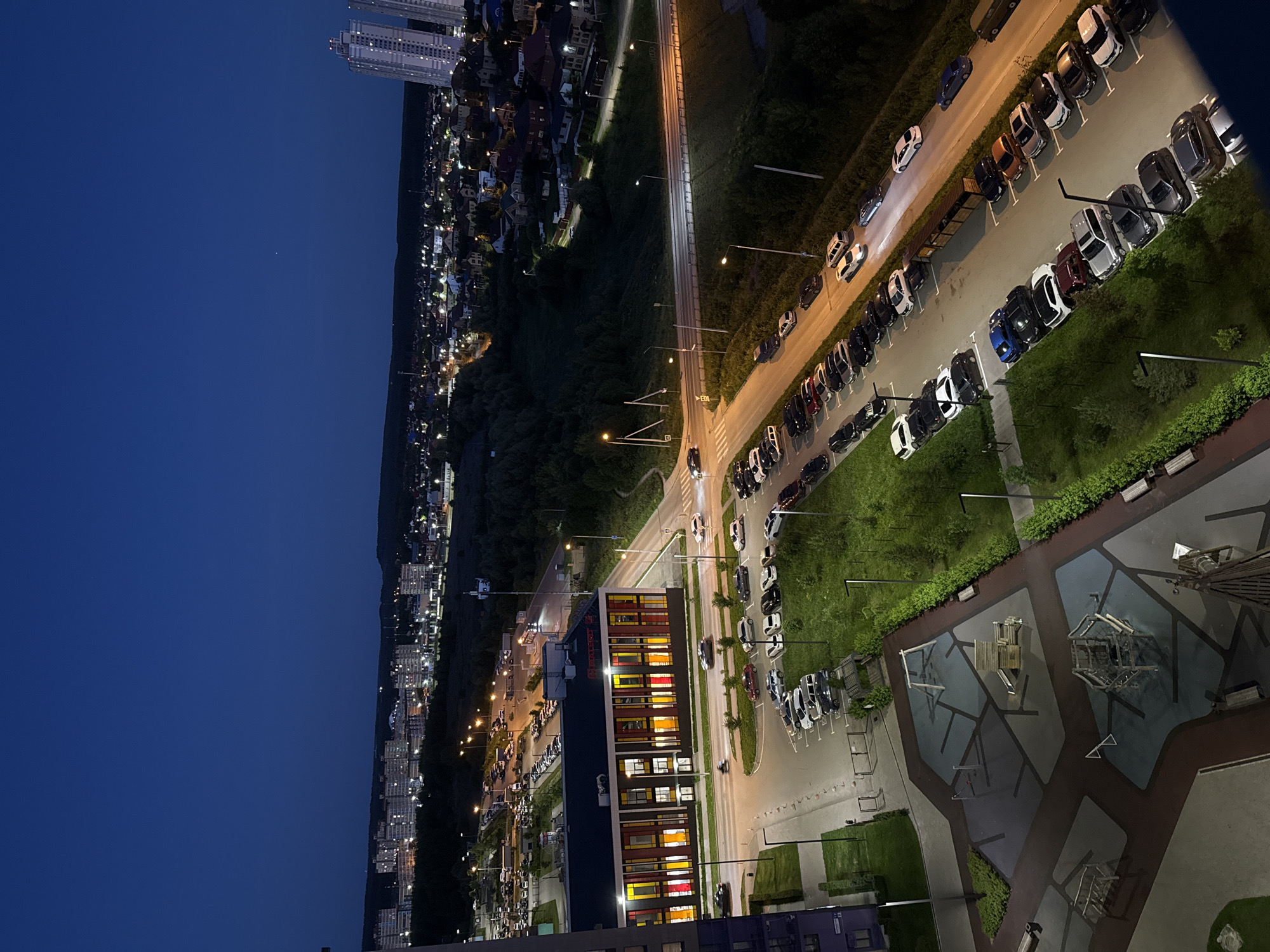 This aerial nighttime view shows a suburban landscape in Perm, Russia, with a focus on a residential area and a street lined with parked cars. The scene is dominated by the deep blue of the twilight sky on the left and the illuminated city on the right. Tall, modern apartment buildings with many lit windows are visible in the distance, suggesting a densely populated area.
In the foreground and midground, a long street is densely packed with parked cars of various colors, including white, black, silver, and red. The street is illuminated by streetlights, casting long shadows and highlighting the parked vehicles. Adjacent to the street is a strip of green grass, and further on, more residential buildings with glowing windows can be seen nestled among trees. A contemporary building with a colorful facade, featuring windows with red, yellow, and white lights, stands out in the midground. A playground with wooden structures is also visible in the lower right portion of the image.
The overall impression is one of a quiet evening in a city, with the lights of homes and vehicles creating a warm glow against the darkening sky. The composition suggests the perspective is from a high vantage point, possibly a tall building. No people are clearly discernible in the image, and no text is visible. The time of day is clearly evening or night, as indicated by the dark sky and illuminated lights. The weather appears calm, with no visible signs of rain or wind.