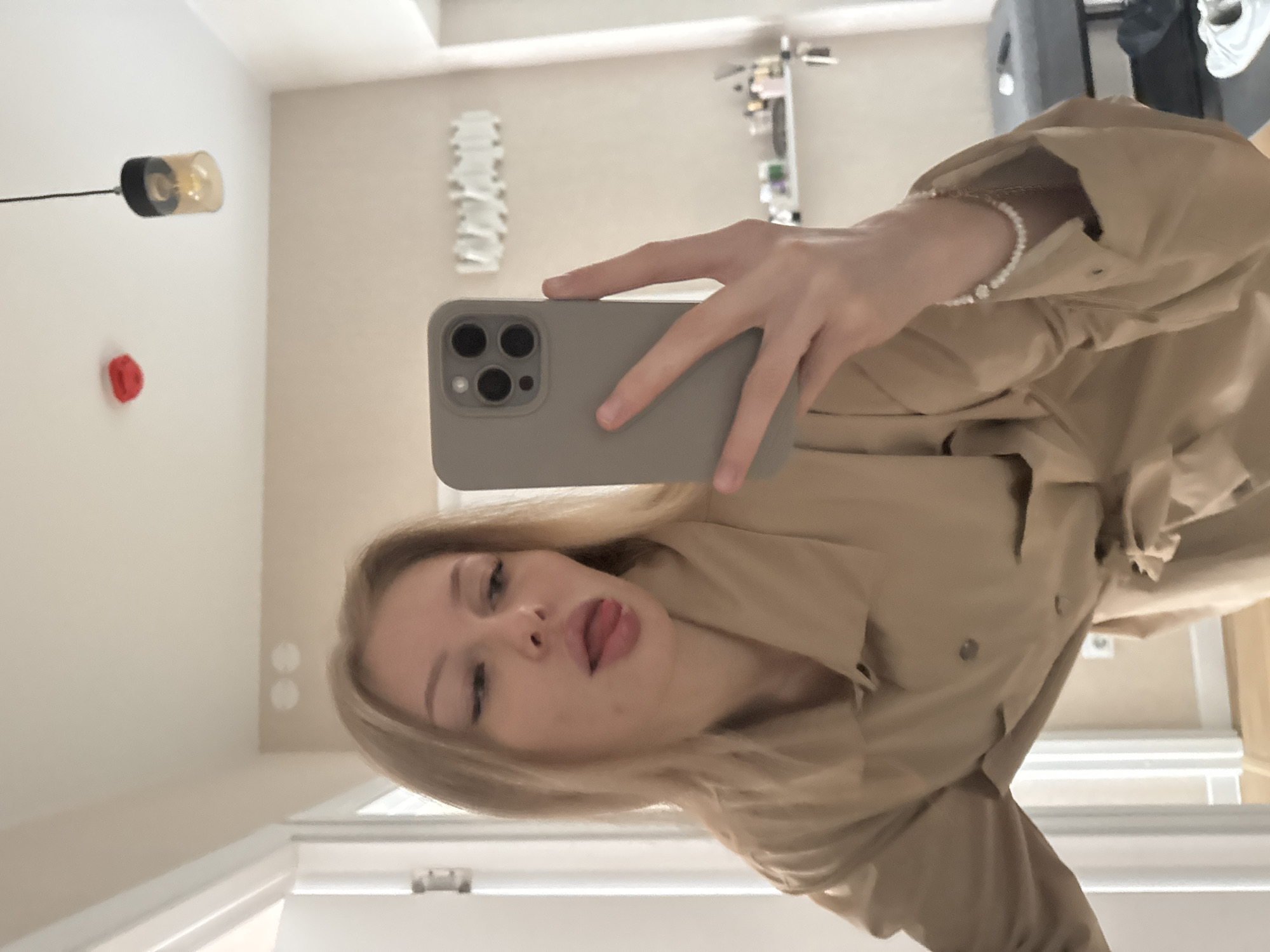 A young, fair-skinned woman with blonde hair is taking a selfie indoors. She is holding a gray smartphone up to her face. She is wearing a light brown jacket and a bracelet. The background appears to be an interior space, possibly a bathroom, with light-colored walls. Details include a light fixture, a white decorative wall piece, a shelf with various items, and a partially visible piece of equipment in the background. The location is Perm, Russia.