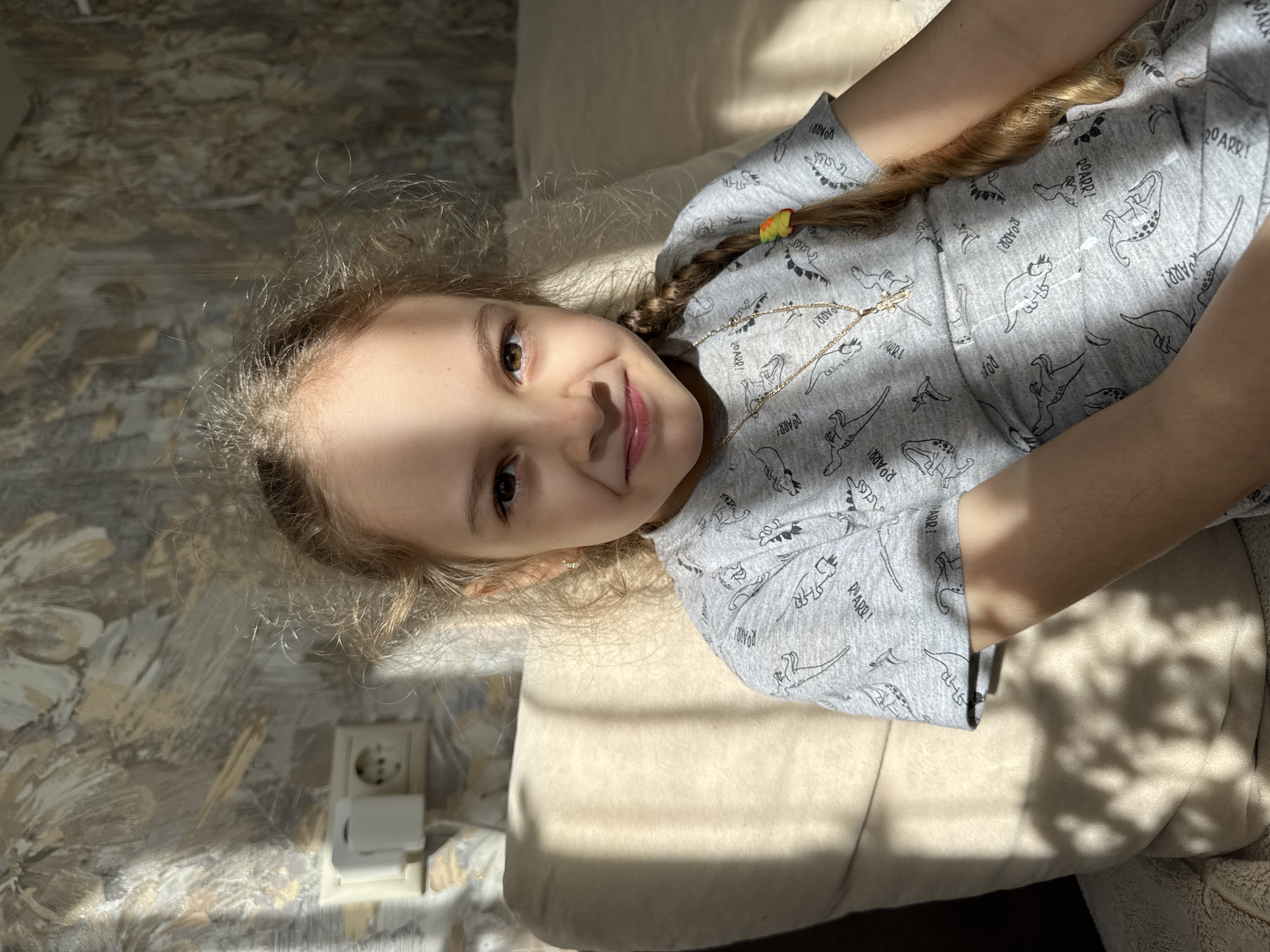 A girl with blonde hair, styled in a braid with a yellow hair tie, is lying on a light beige sofa. She is wearing a grey t-shirt with dinosaur prints and a gold necklace with a cross pendant. A stripe of sunlight crosses her face, creating high contrast. The background includes a wall with floral-patterned wallpaper and an electrical outlet. The location is unknown.