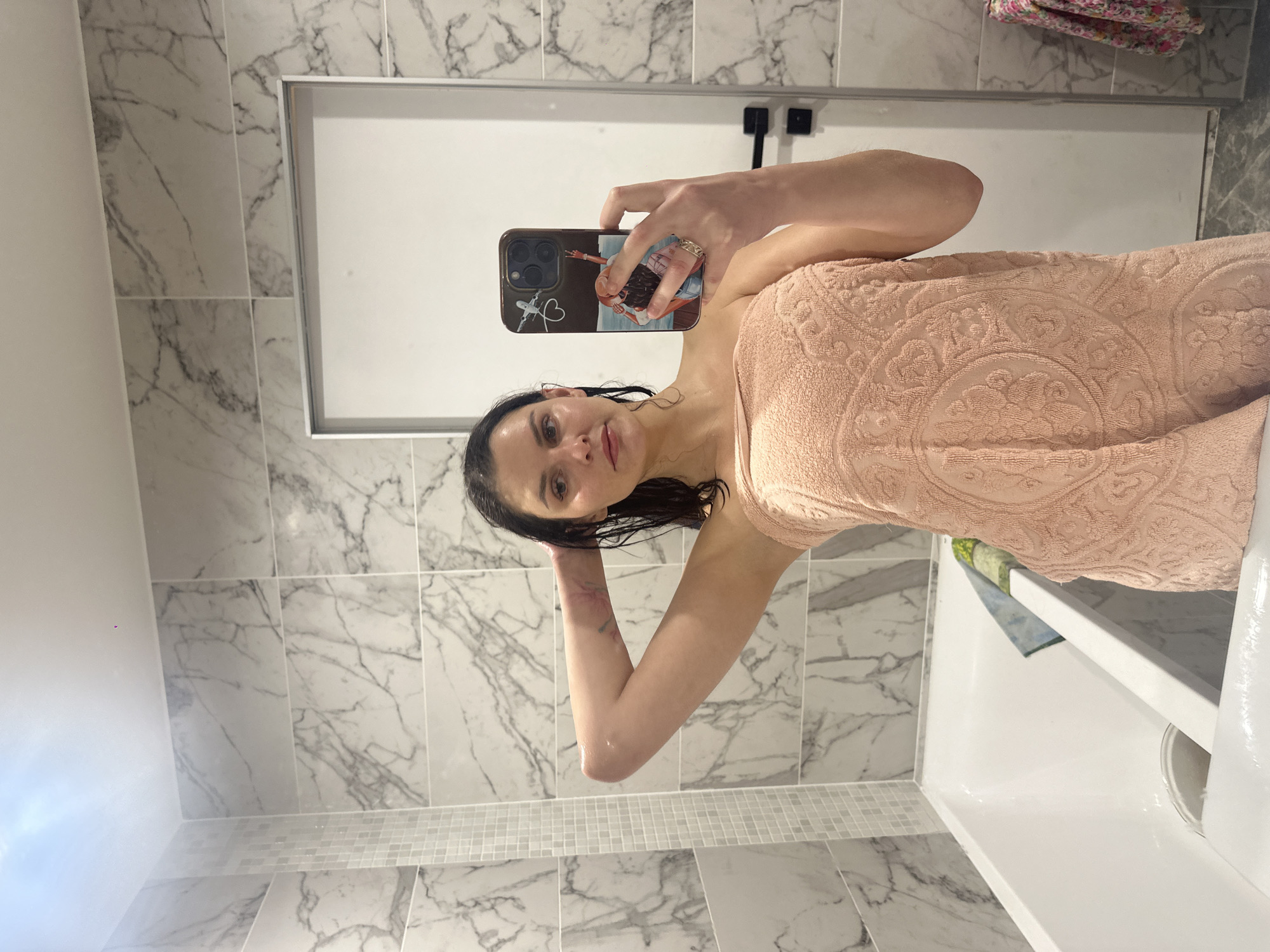 In an indoor bathroom with marble tile walls and a white bathtub, a woman wrapped in a peach-colored towel stands and holds her phone up to take a selfie in the mirror. Her hair is dark and wet, and her makeup is light. She is wearing a ring on her left hand, which is visible in the mirror as she holds her phone. The phone case features an illustration of two women on a boat.