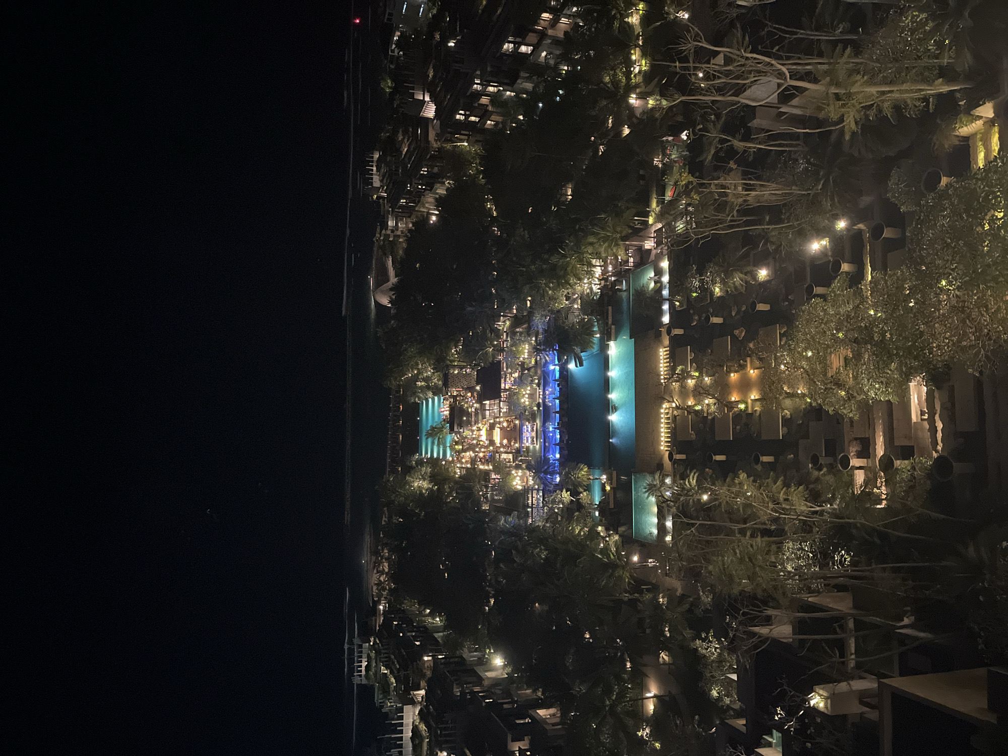 This is a nighttime aerial view of a resort in Kuta Selatan, Indonesia. The resort features multiple illuminated swimming pools, some with a vibrant turquoise glow, others with a softer blue light. Numerous palm trees and other lush vegetation are scattered throughout the property, illuminated by warm-toned lights. Buildings with lit windows are visible, suggesting occupied rooms or common areas. In the distance, the ocean is visible, with a faint horizon line under a dark sky. The overall impression is of a well-lit, luxurious resort complex at night.