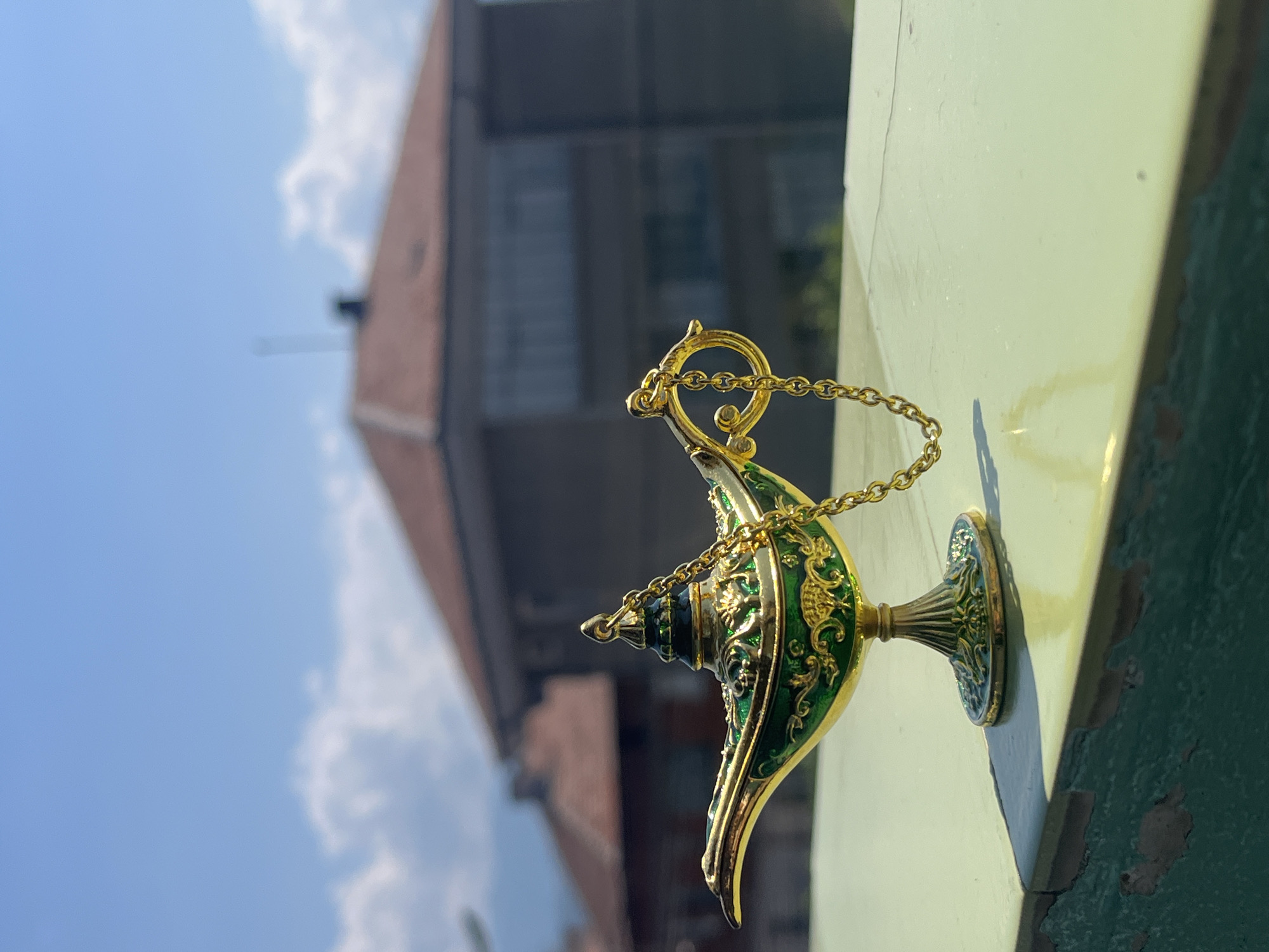 This is a close-up, vertically oriented outdoor photograph of a decorative, golden Aladdin-style lamp with green enamel accents. The lamp is placed on a pale green surface, casting a distinct shadow. A thin gold chain is attached to the top of the lamp and drapes over its side.
In the background, out of focus, are blurred architectural elements of buildings with reddish-brown roofs, suggesting an urban or suburban setting. The sky above is a vibrant blue with scattered white clouds, indicating fair weather during the daytime. The lighting suggests it is likely morning or late afternoon, given the angle of the sunlight creating the shadow.
The object's ornate design, with intricate patterns on the green enamel and gold detailing, suggests it is a decorative item, possibly a souvenir or collectible. There is no discernible text in the image. The overall mood is peaceful and serene, with the focus sharply on the lamp, contrasted against the soft blur of the background. The location context of Pokrovnik, Bulgaria, provides a geographical anchor to the scene.