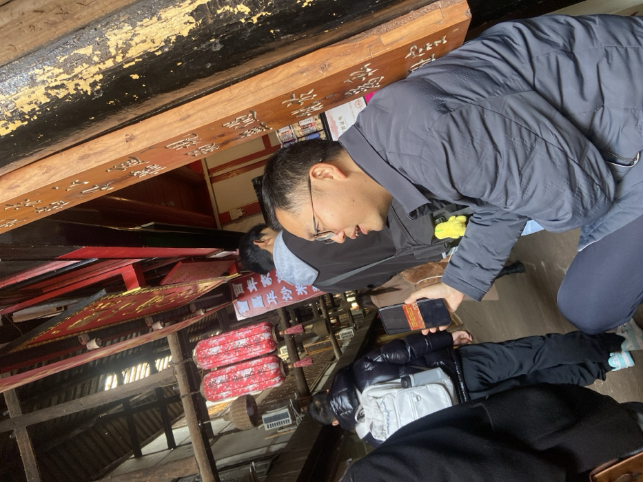 A man with glasses wearing a gray jacket looks at his phone. Several people surround him, all indoors. Red lanterns and wooden structures adorn the setting, suggesting a temple or traditional building. Carved wooden beams with Chinese characters run along the ceiling. The angle of the image is slightly tilted.