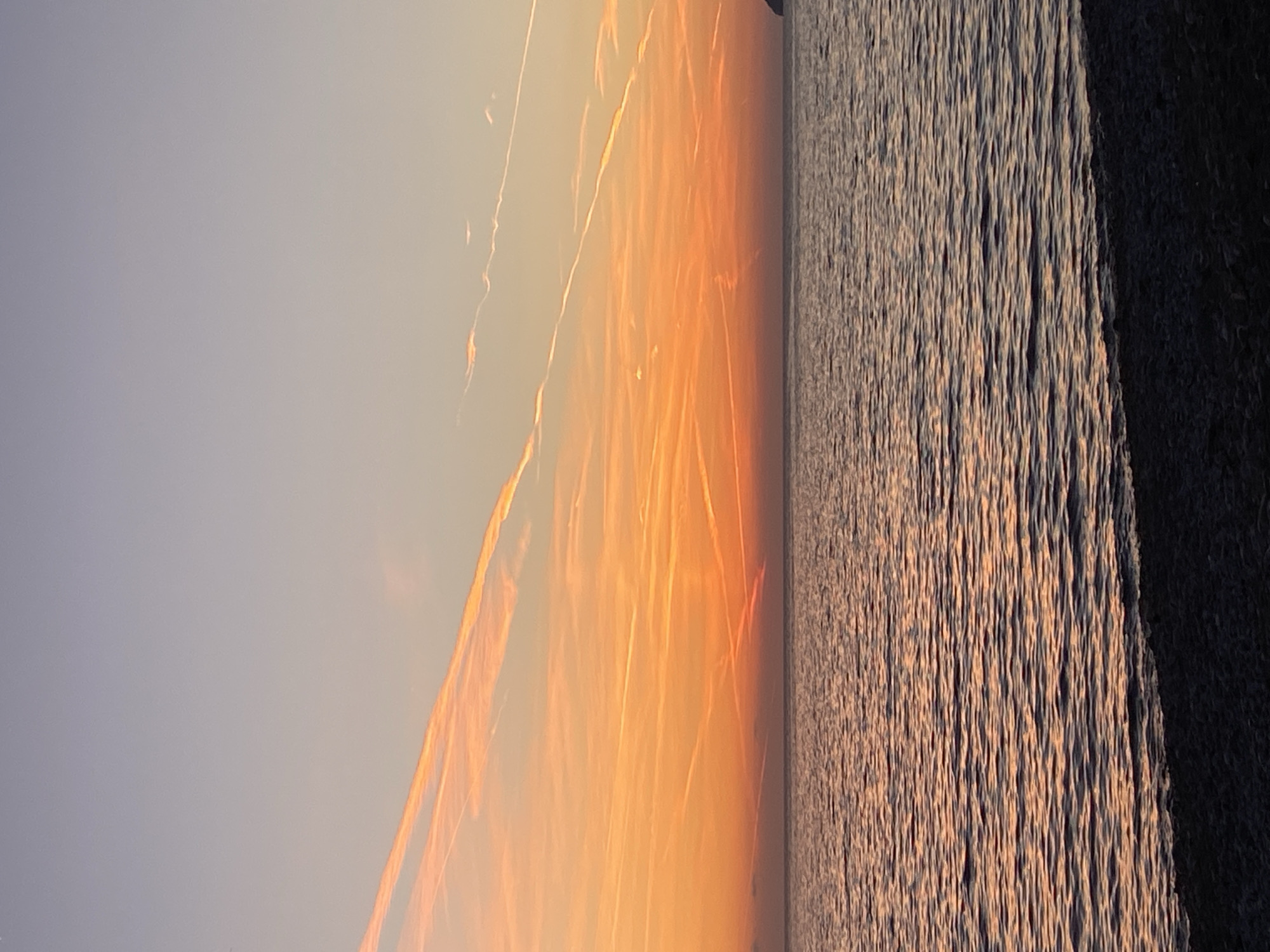 A sunset view from Bar, Montenegro shows a body of water adjacent to a dark shoreline, possibly a breakwater or rocky coast. The sun is setting, casting a deep orange hue across the sky, with contrails left by airplanes crisscrossing the upper portion of the frame. The water reflects the golden light of the sunset, creating shimmering patterns on the surface. The sky appears light blue in the areas not directly impacted by the sun's glow.