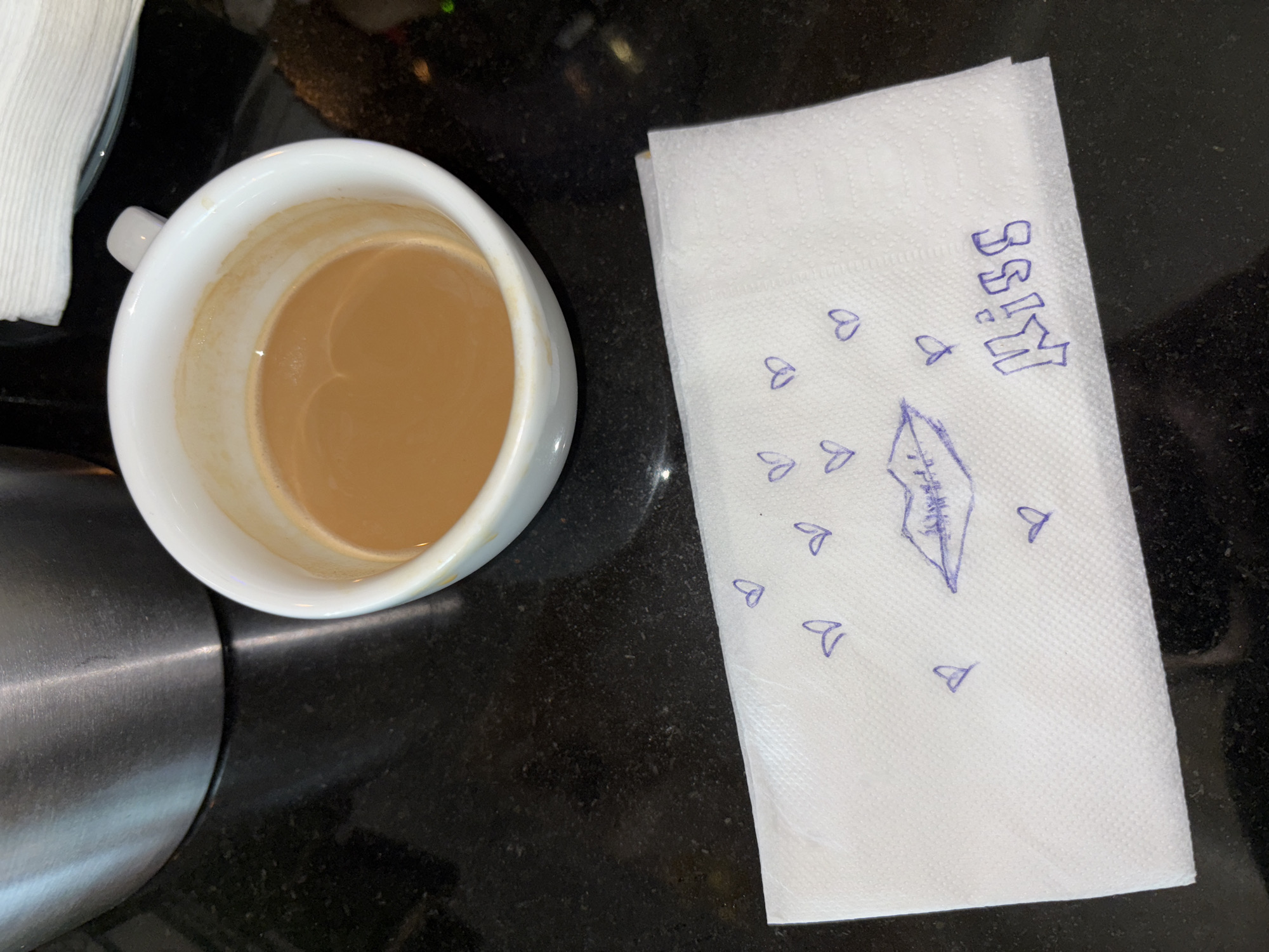 The scene shows a white coffee cup filled with coffee next to a white paper napkin on a black surface. The napkin has the word "KISS" written in blue ink, along with a drawing of lips and several small heart shapes. A part of a metal object is visible on the left side of the frame. The location cannot be determined as Sofia, Bulgaria, based on the visual evidence.
