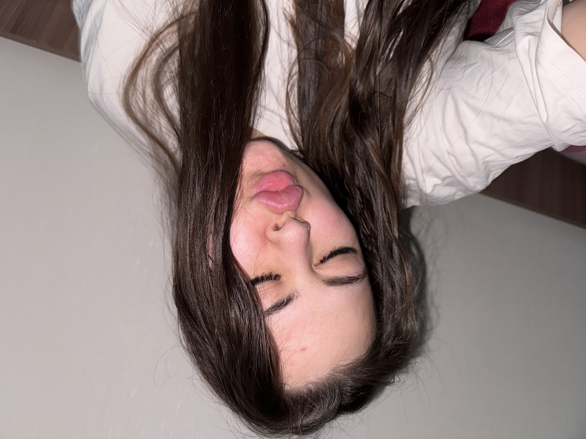 An informal, close-up portrait shows a young woman lying down in an indoor setting in Saint Petersburg, Russia. Her head is angled, and her long, dark brown hair is spread out around her. She is wearing a light-colored, possibly white, long-sleeved shirt. The woman is making a playful or humorous expression, with her tongue slightly extended and her eyes closed or squinted. A small nose ring is visible on her right nostril, and a minor blemish appears on her forehead. The background features a plain, light-colored wall and some dark brown furniture or surfaces, suggesting a casual home environment. The lighting is soft, likely artificial, creating a candid atmosphere. No visible text is present.