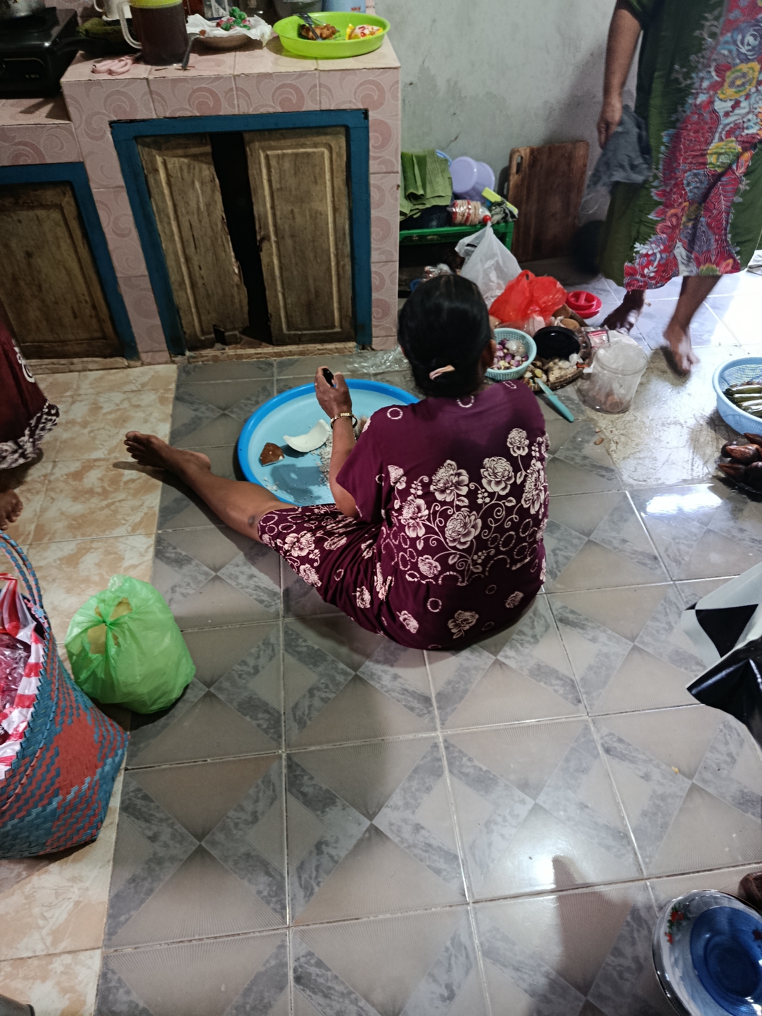 This image depicts an indoor scene, likely a kitchen or food preparation area in Sarwadadi, Indonesia. A woman is seated on the floor, facing away from the camera, dressed in a dark purple dress with a floral pattern. She is holding a utensil, possibly a knife, and is engaged with a blue plastic basin in front of her, which contains pieces of coconut. Beside the basin is a small pile of what appears to be chopped ingredients.
The environment shows a tiled floor with a geometric pattern. To the left of the woman is a large, woven basket with red and blue accents, and a green plastic bag. In the background, there are kitchen cabinets with wooden doors, and a countertop holding various items including containers, a mug, and a plate of food. Another person, only their lower legs and feet are visible, is standing in the upper right portion of the image, wearing a patterned skirt. The overall impression is one of a domestic setting where food is being prepared.