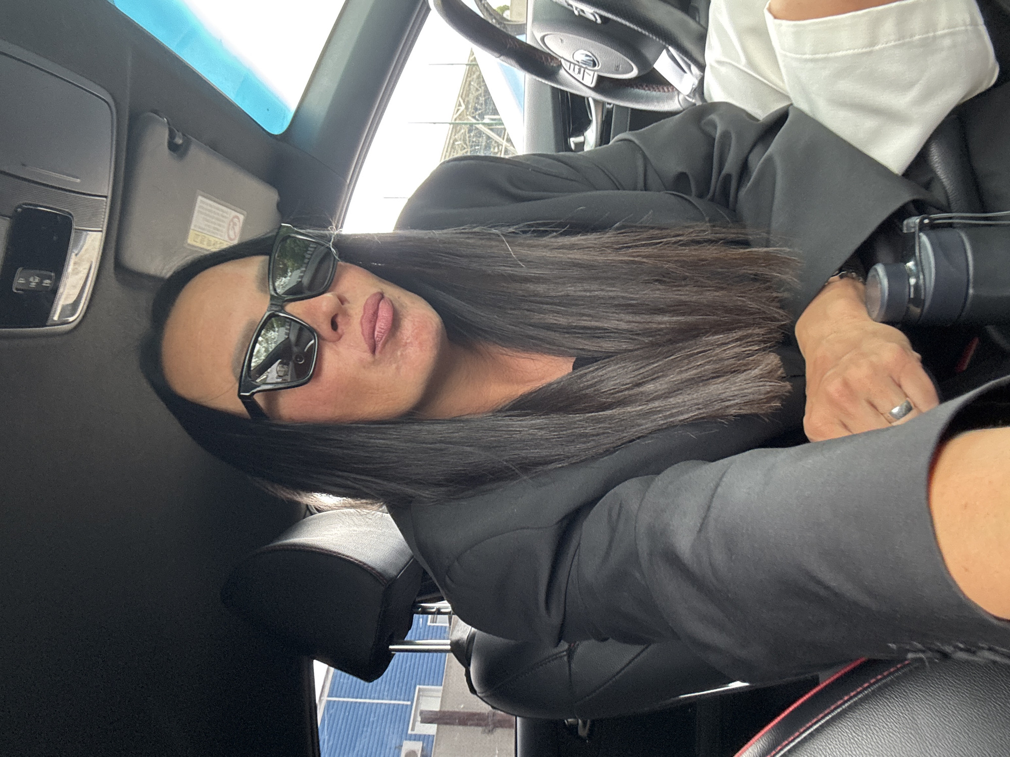 A medium shot captures a person seated in the driver's seat of a car, wearing a black blazer, white shorts, and black sunglasses. The person's long, dark hair falls forward, partially obscuring their face. They are looking down and slightly to the left, with their lips pursed. A silver ring is visible on their left hand, which rests on a dark-colored water bottle. The car's interior is visible, including the steering wheel and part of the passenger seat. Outside the car, a blue building and some structures can be seen. The lighting suggests it is daytime. The specific location in Chaikovsky, Russia, cannot be determined from the visual information provided.