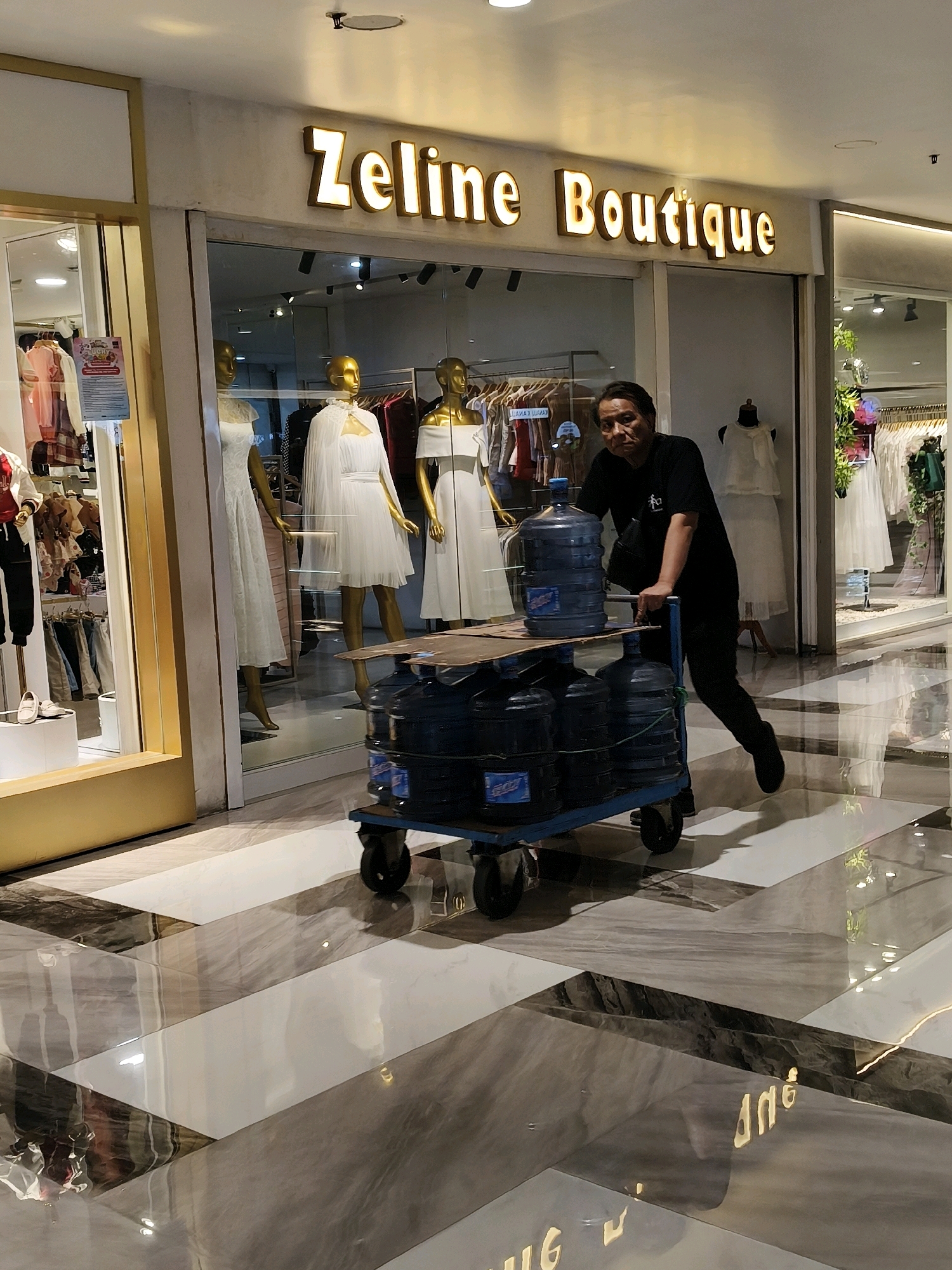 A man pushes a dolly loaded with several large blue water jugs down a polished floor in a shopping mall. The dolly has a blue frame and black wheels. The water jugs are clear plastic with blue labels. The man is wearing a black t-shirt with a white symbol on it and black pants. He is pushing the dolly from the right side. Behind him, there is a storefront with the illuminated sign "Zeline Boutique". The boutique displays mannequins dressed in white clothing. The floor is made of large, diagonally placed tiles in shades of gray, white, and black with a high-gloss finish, reflecting the lights of the mall. The overall environment appears to be a modern indoor shopping mall located in Surabaya, Indonesia.