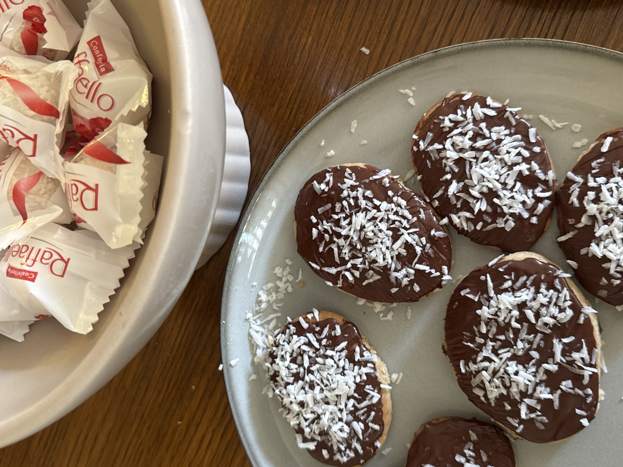 The image shows a wooden table with a light gray bowl of individually wrapped Raffaelo confections to the left. To the right, on a light gray plate, are six oval-shaped cookies or pastries covered in chocolate and sprinkled with shredded coconut. The scene does not provide information to identify its location as Burgas, Bulgaria or any other specific location.