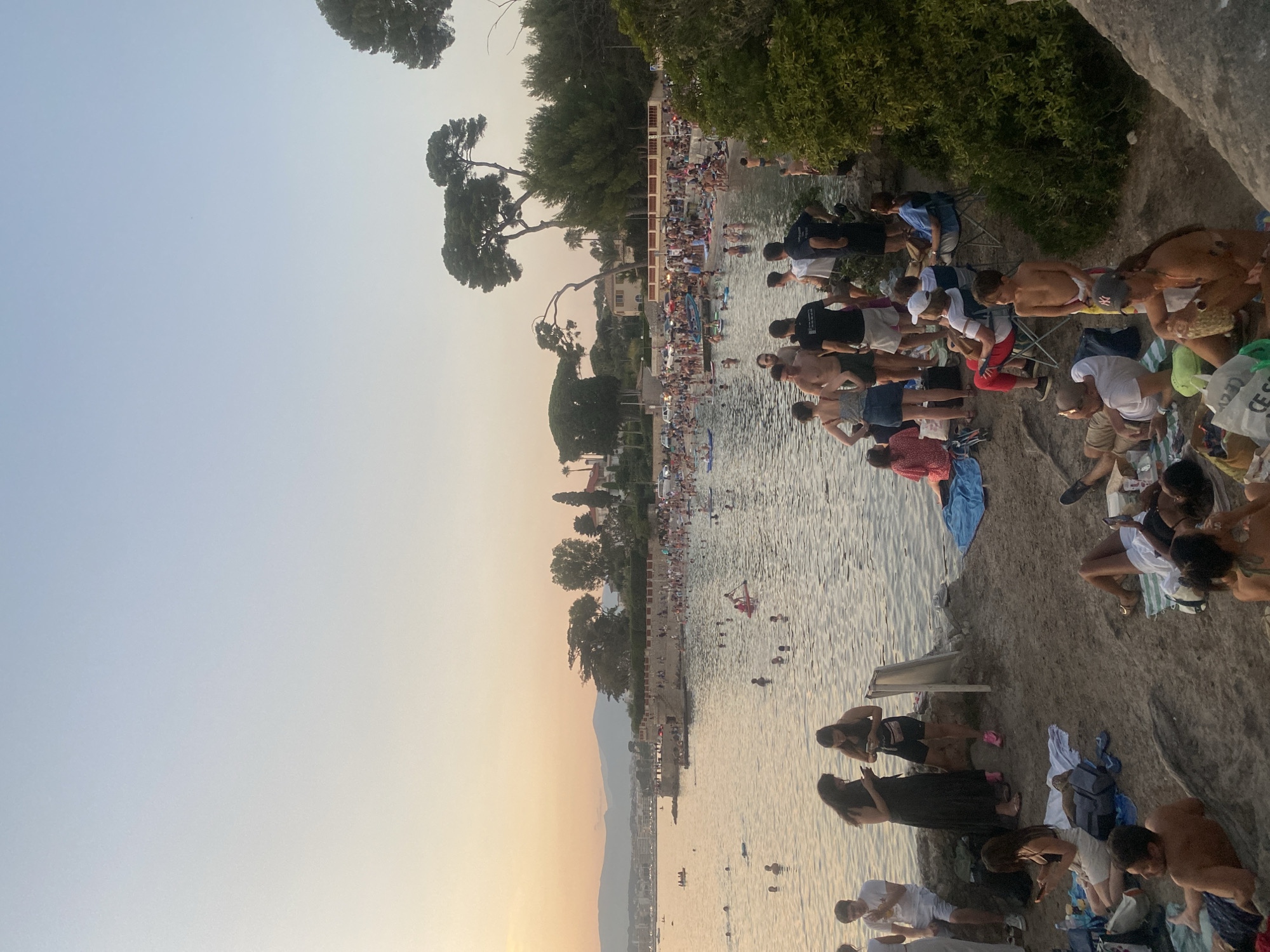 This is a medium shot, taken from a slightly elevated position, of a crowded beach scene in Antibes, France, during the late afternoon or early evening. The sky is a pale gradient of blue and orange, indicating sunset. The water of the Mediterranean Sea is calm and reflects the warm light.
In the foreground, on the rocky shore, several groups of people are relaxing on towels and blankets. Some are sitting, some are lying down, and many are looking at their phones. One person is holding a pineapple. There are also bags and other personal belongings scattered around.
Further back, the water is filled with people swimming, wading, and on paddleboards. A long, stone wall with a railing runs along the edge of the water in the mid-ground, with many people standing and sitting on it, overlooking the sea. Beyond the wall, the beach is densely populated with sunbathers and swimmers. Buildings and trees are visible on the hillside behind the beach. In the distance, faint mountains can be seen against the horizon. The overall impression is one of a lively and popular public space enjoying the evening.