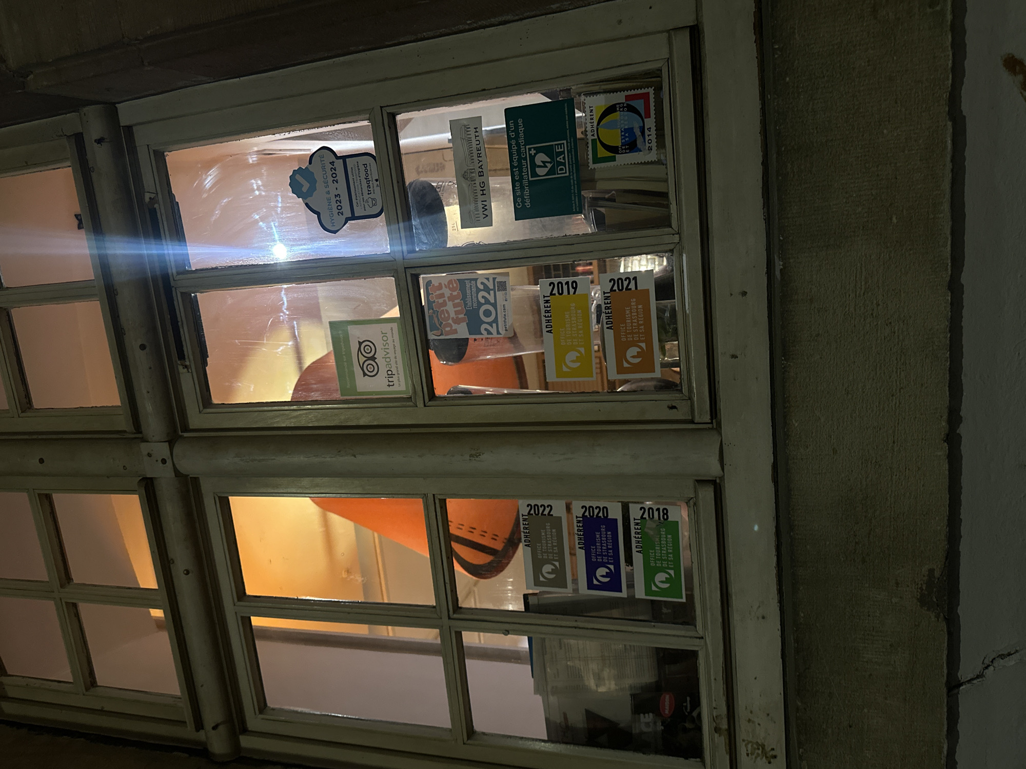 A window in Strasbourg, France, is visible from the exterior. The window is divided into many panes, and several stickers and labels are affixed to the glass. "Hygiene & Security 2023-2024" is present, along with a TripAdvisor logo. A "Petit Futé 2022" sticker is visible. There is a sticker that says "VWI HG BAYREUTH" and another one indicating the presence of a defibrillator ("DAE"). There are also stickers indicating the years 2014, 2018, 2019, 2020, 2021, and 2022, and the word "ADHÉRENT" is featured on the stickers, along with the text "OFFICE DE TOURISME DE STRASBOURG ET SA REGION." Through the window, there is an orange cone and an interior space. The light suggests that it is nighttime. The exterior wall is a rough grey.