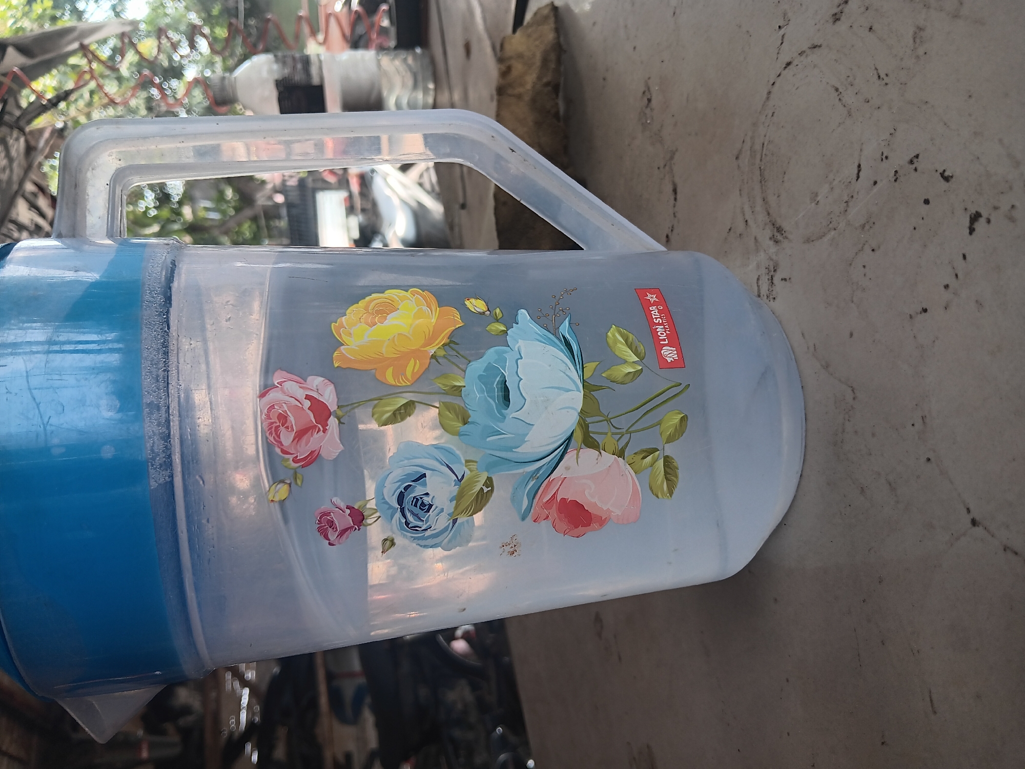 A clear plastic jug with a blue lid is filled with water and decorated with floral motifs, including pink, blue, and yellow roses. A red and white "LION STAR PLASTICS" logo is visible on the side of the jug. The jug rests on a textured, weathered surface, possibly a table, which is littered with small specks of dirt and smudges. In the background, blurry elements suggest an outdoor setting with trees and possibly a vehicle or structure. The lighting indicates it is daytime.